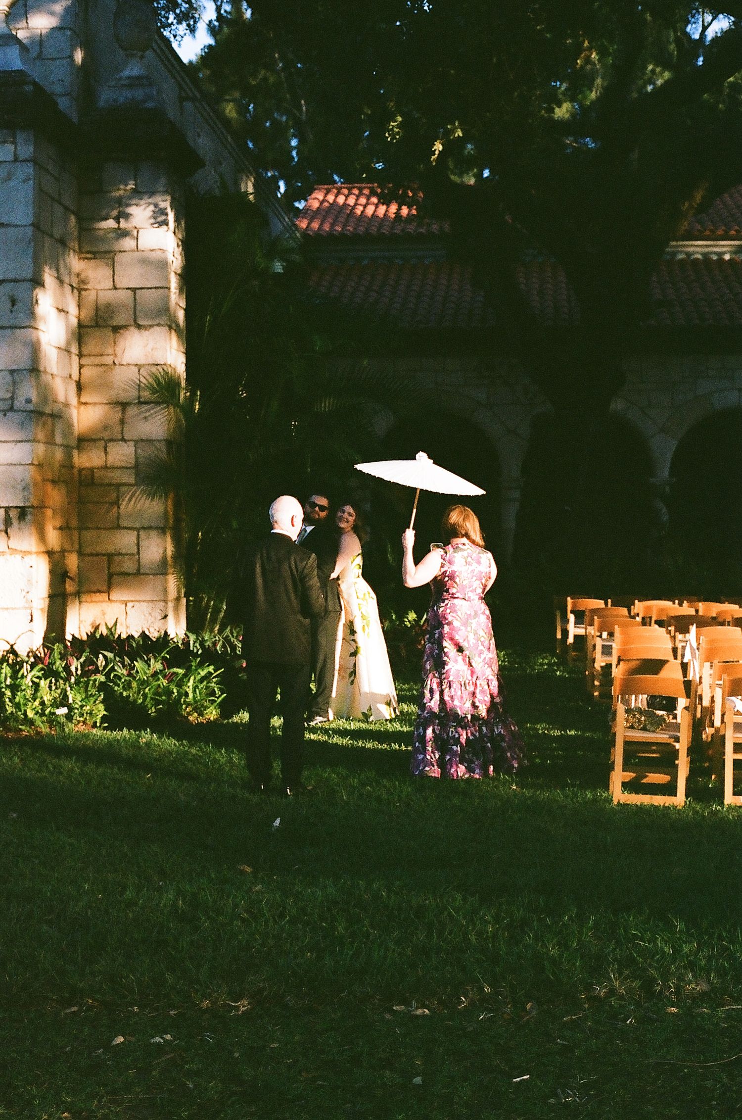 Bride with white parasol walks toward stone chapel with wedding guests seated in wooden chairs.