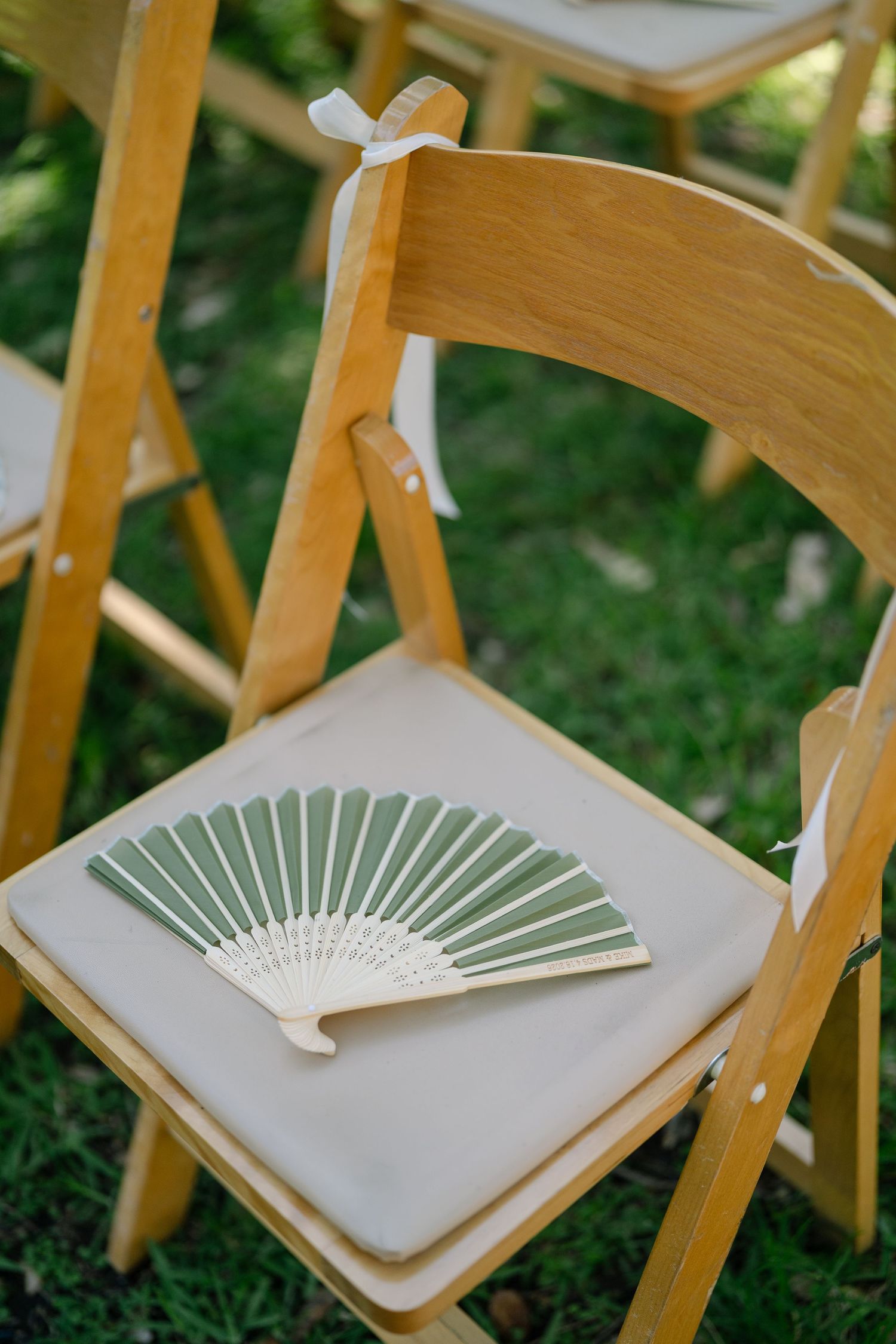 A sage green paper hand fan rests on a white cushioned wooden folding chair at an outdoor wedding ceremony.