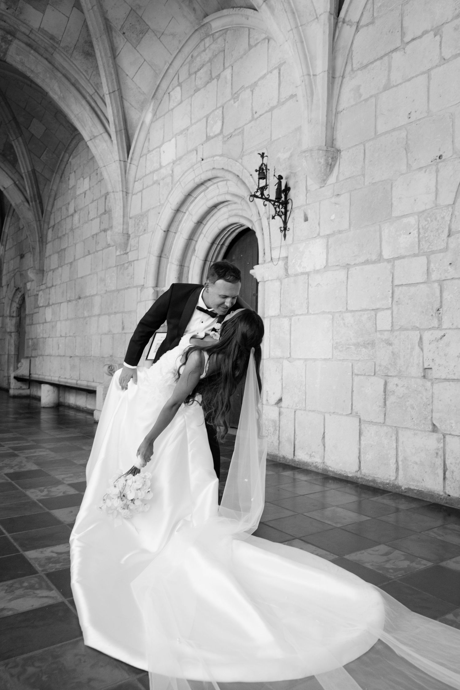 Black and white photo of groom dipping bride in flowing veil inside stone arched wedding venue.