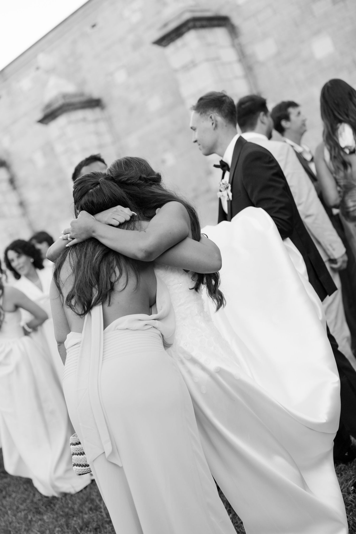 Black and white photo of a bride embracing a bridesmaid in a white jumpsuit at an outdoor wedding venue.