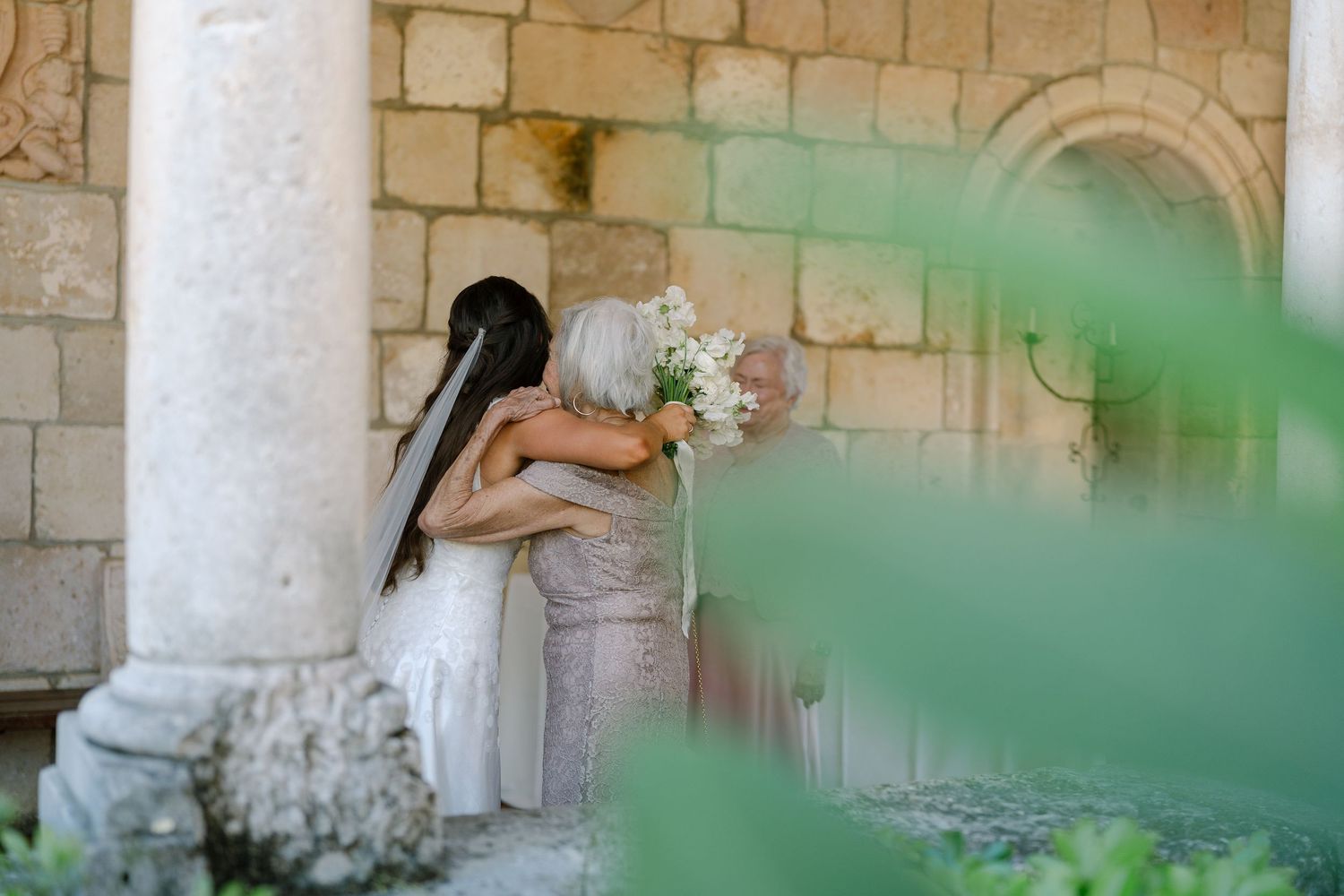 Bride and older woman embrace near stone columns holding white floral bouquet outdoors.