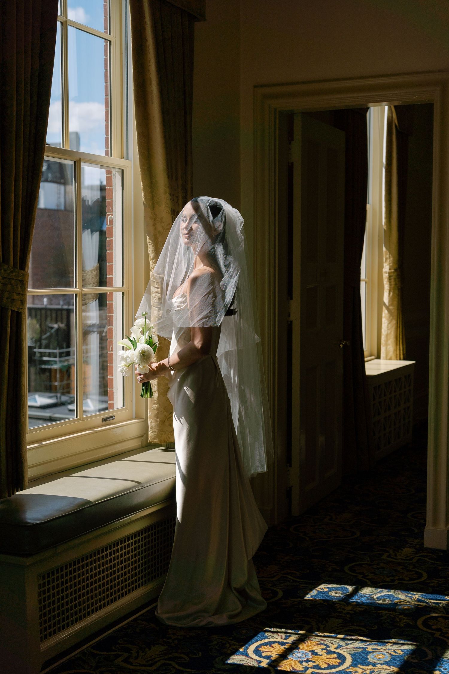 Bride in satin gown and veil stands by sunlit window holding white bouquet before wedding ceremony.