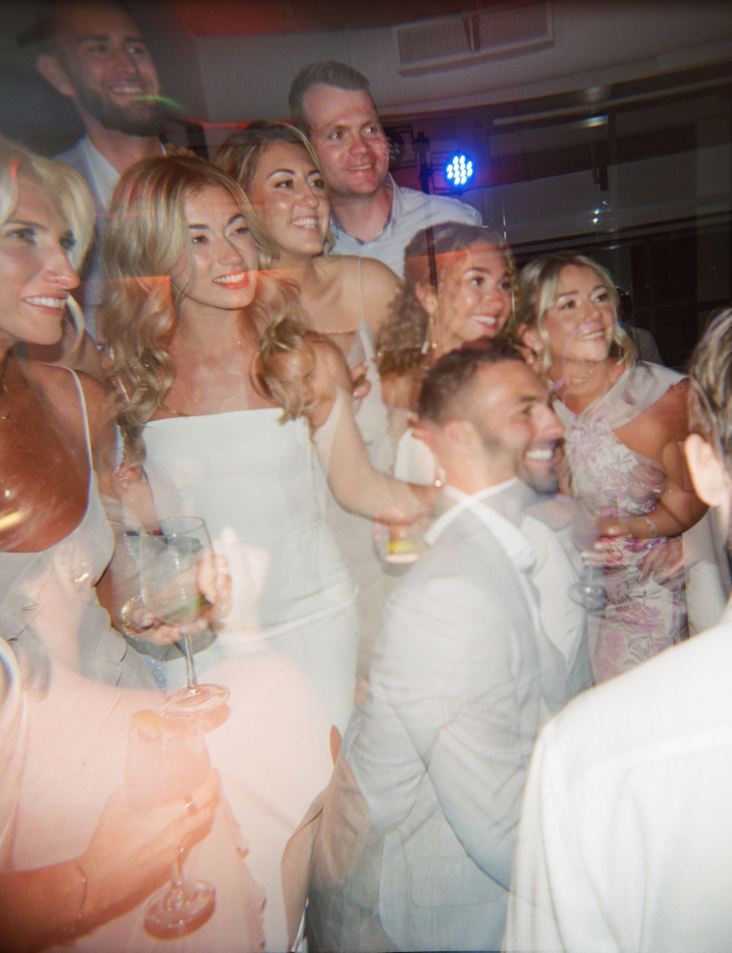 Wedding reception party scene with guests in white and formal attire dancing under ambient lighting.