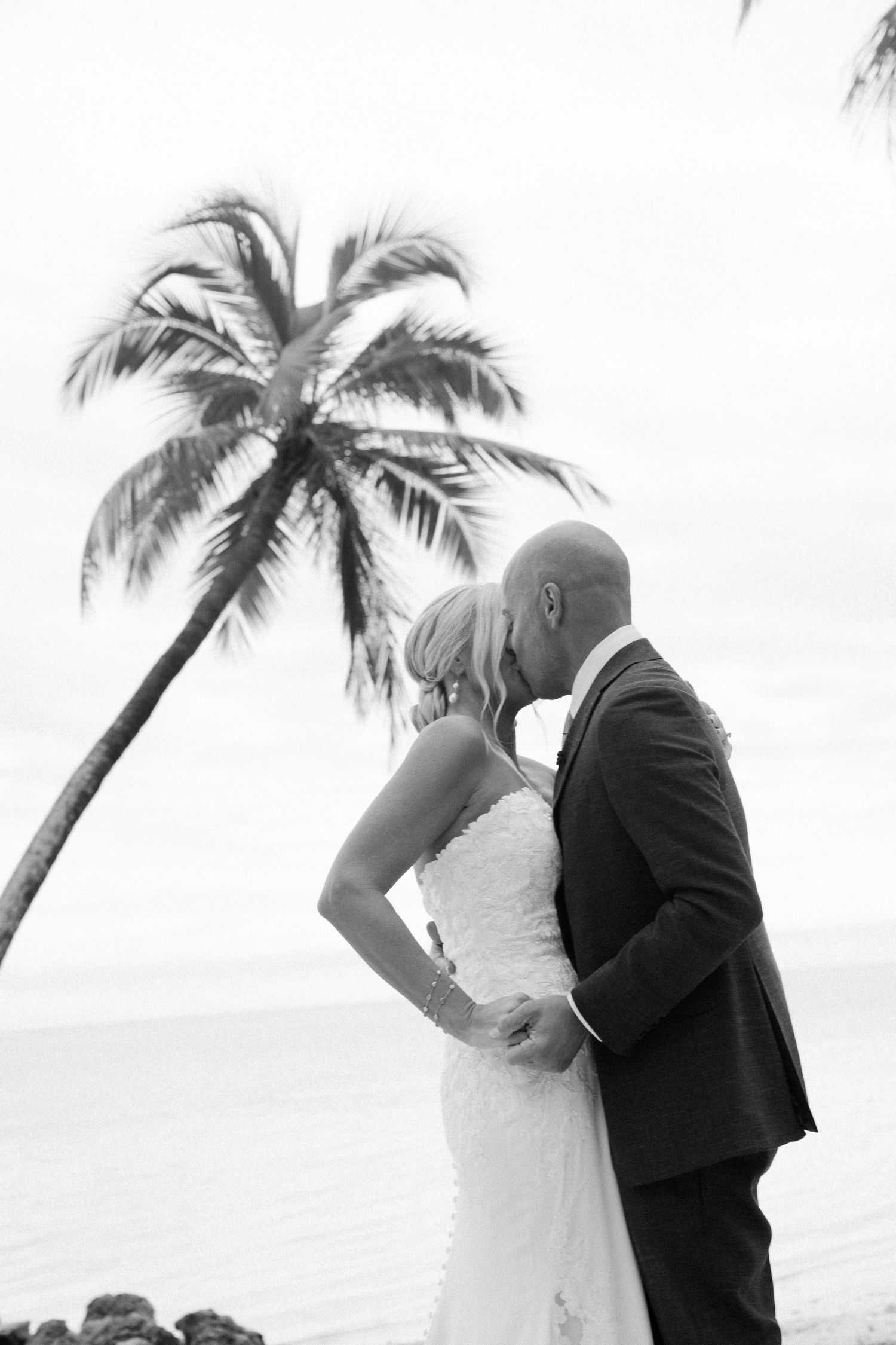 A romantic black and white wedding photo of a couple kissing under a palm tree by the ocean.
