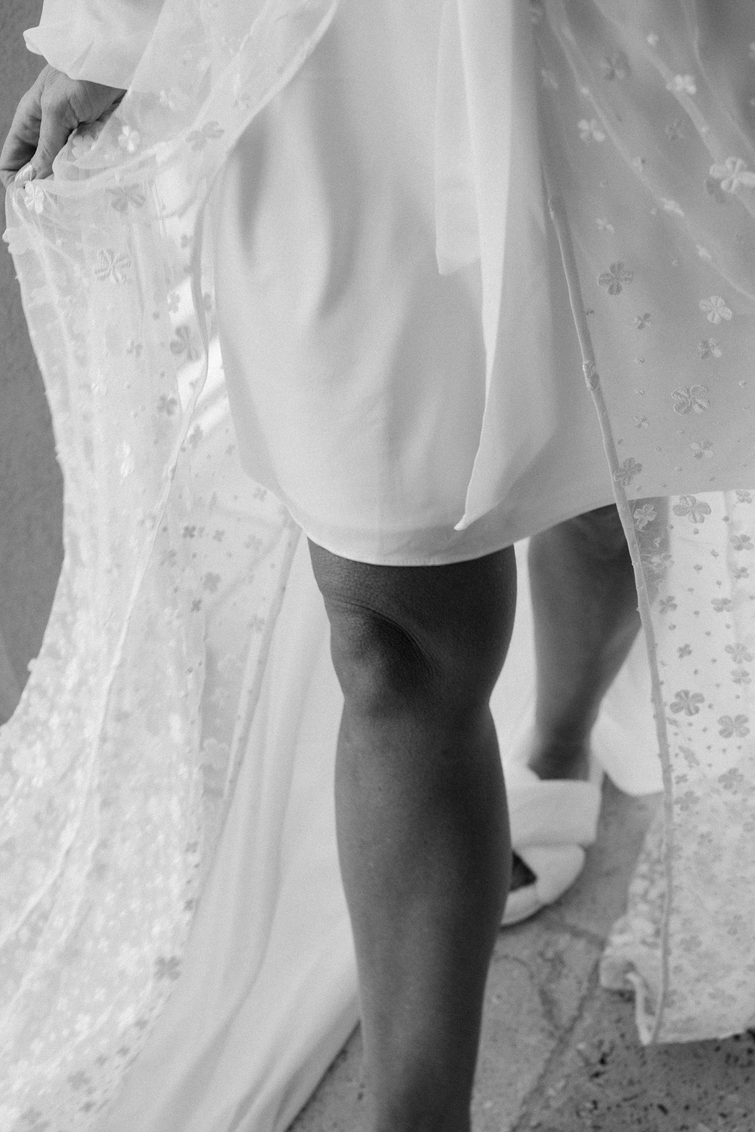 Black and white close-up of legs in white dress and veil fabric showing delicate wedding dress details.