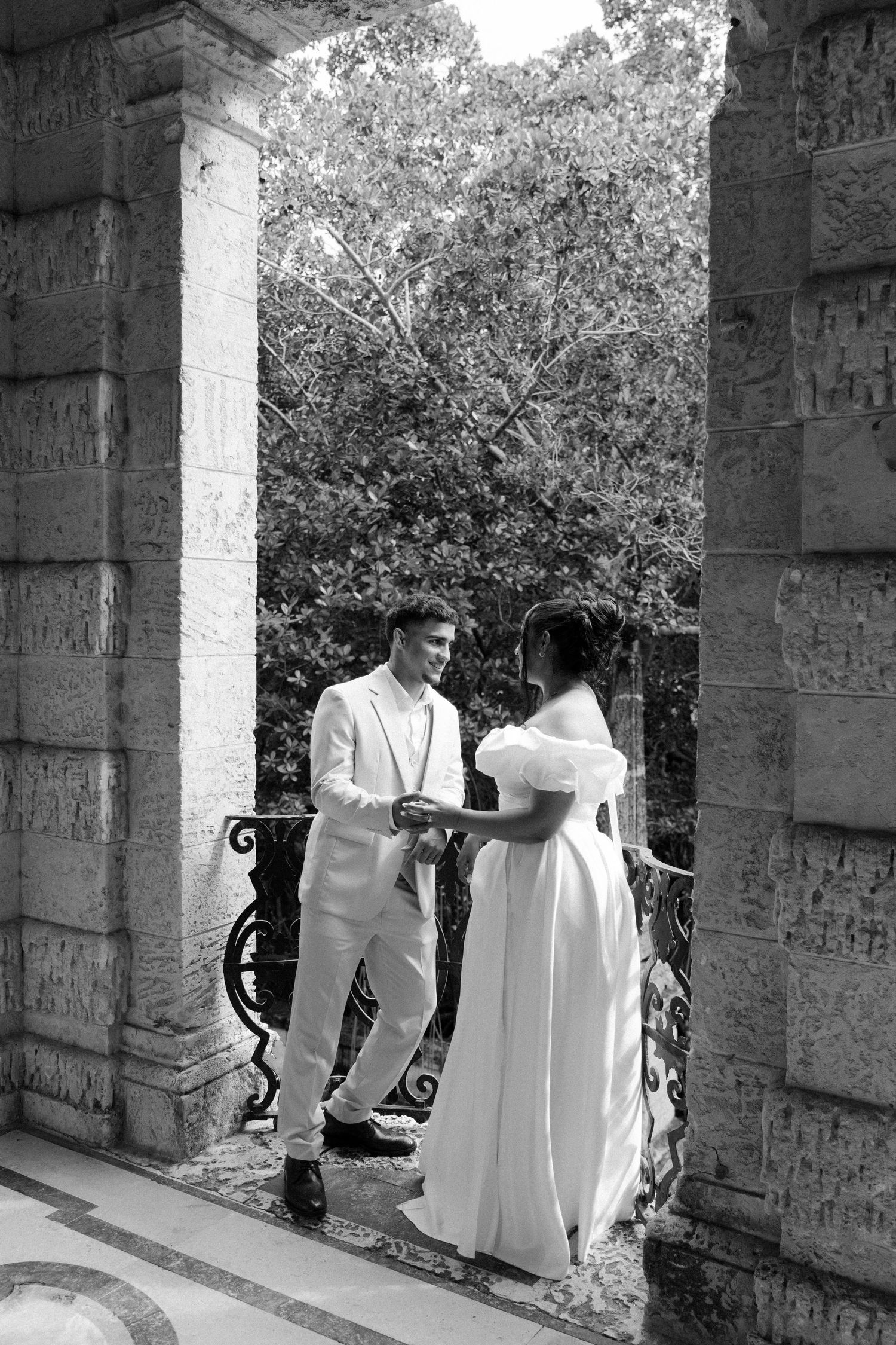 A couple in elegant white attire stand together in a stone archway during what appears to be a wedding photoshoot.