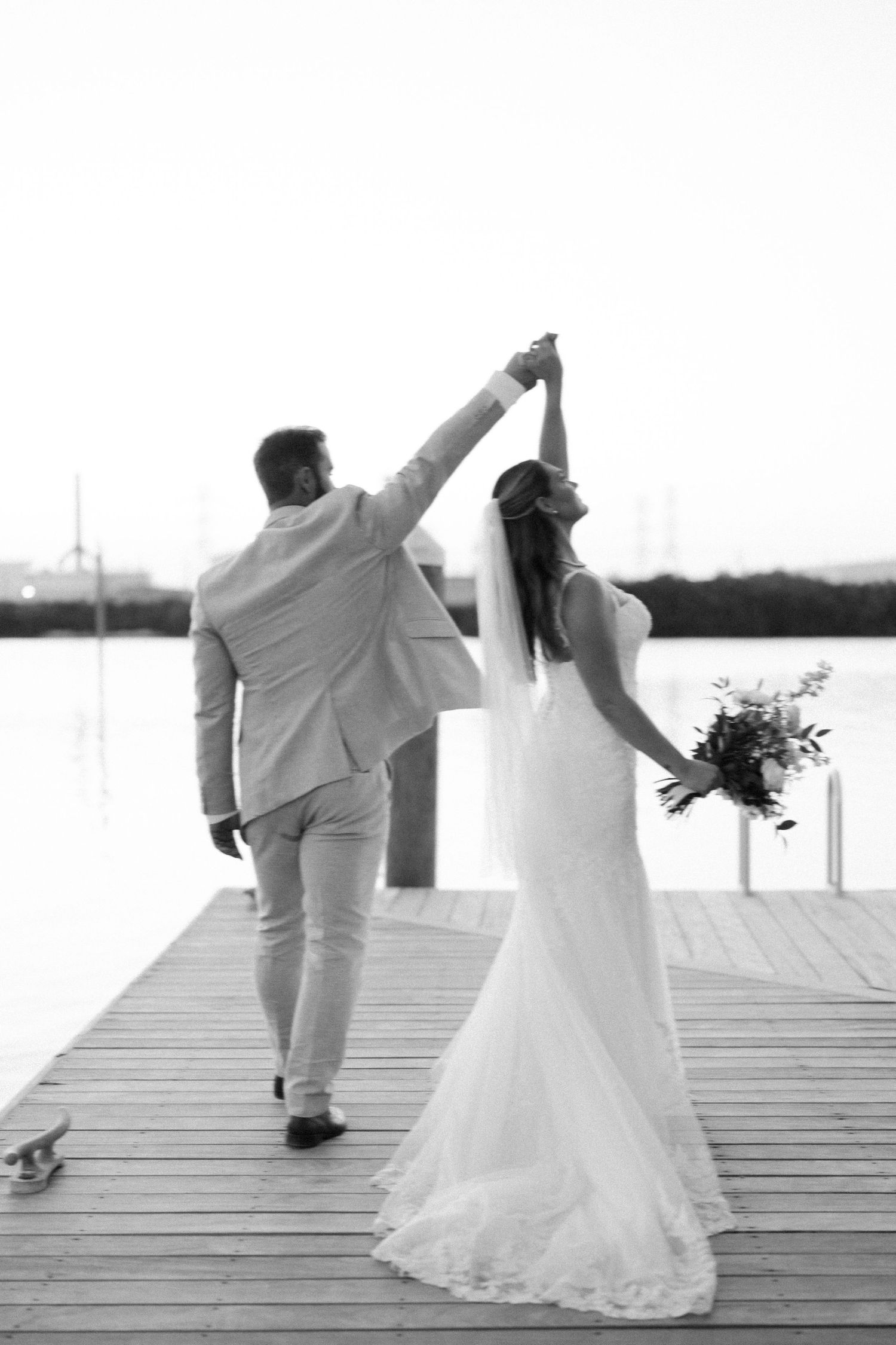 A couple in wedding attire dancing on a wooden pier by the water.
