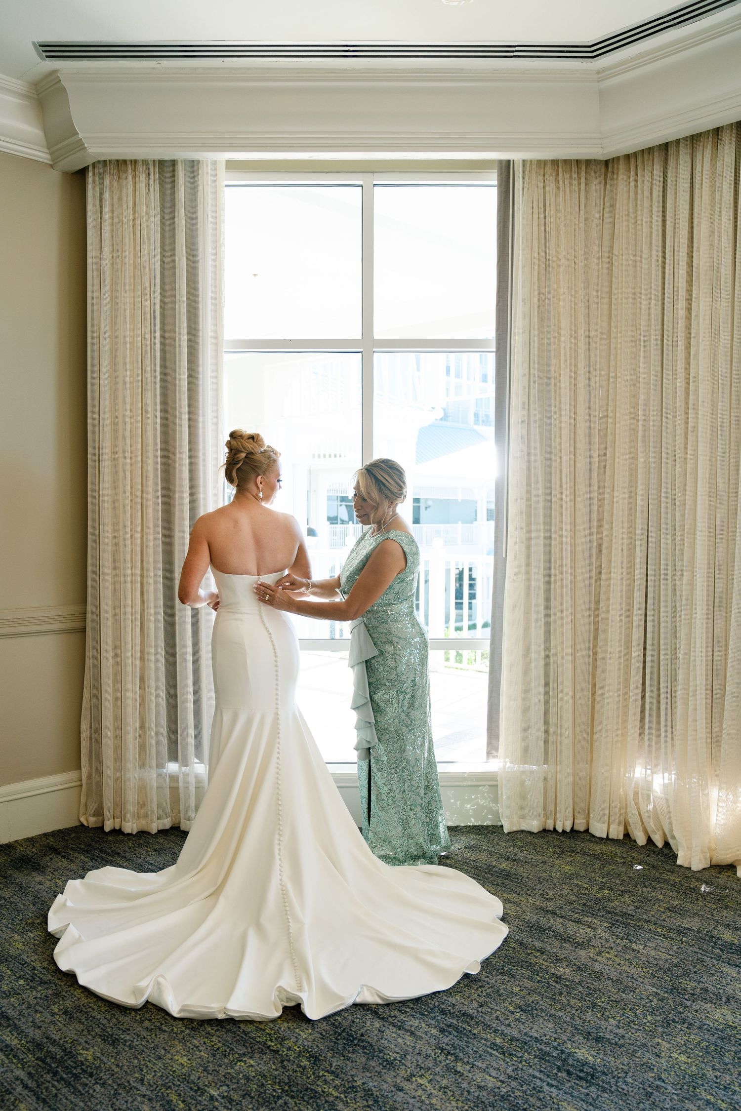 A bride in a flowing white gown gets ready in an elegant hotel room with floor-to-ceiling windows.