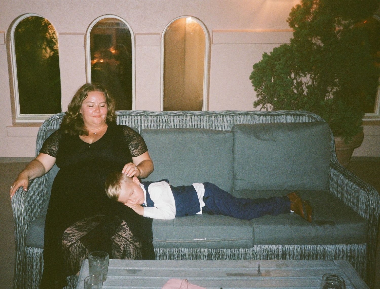 Two people relaxing on a grey outdoor couch at dusk with arched windows visible in the background.
