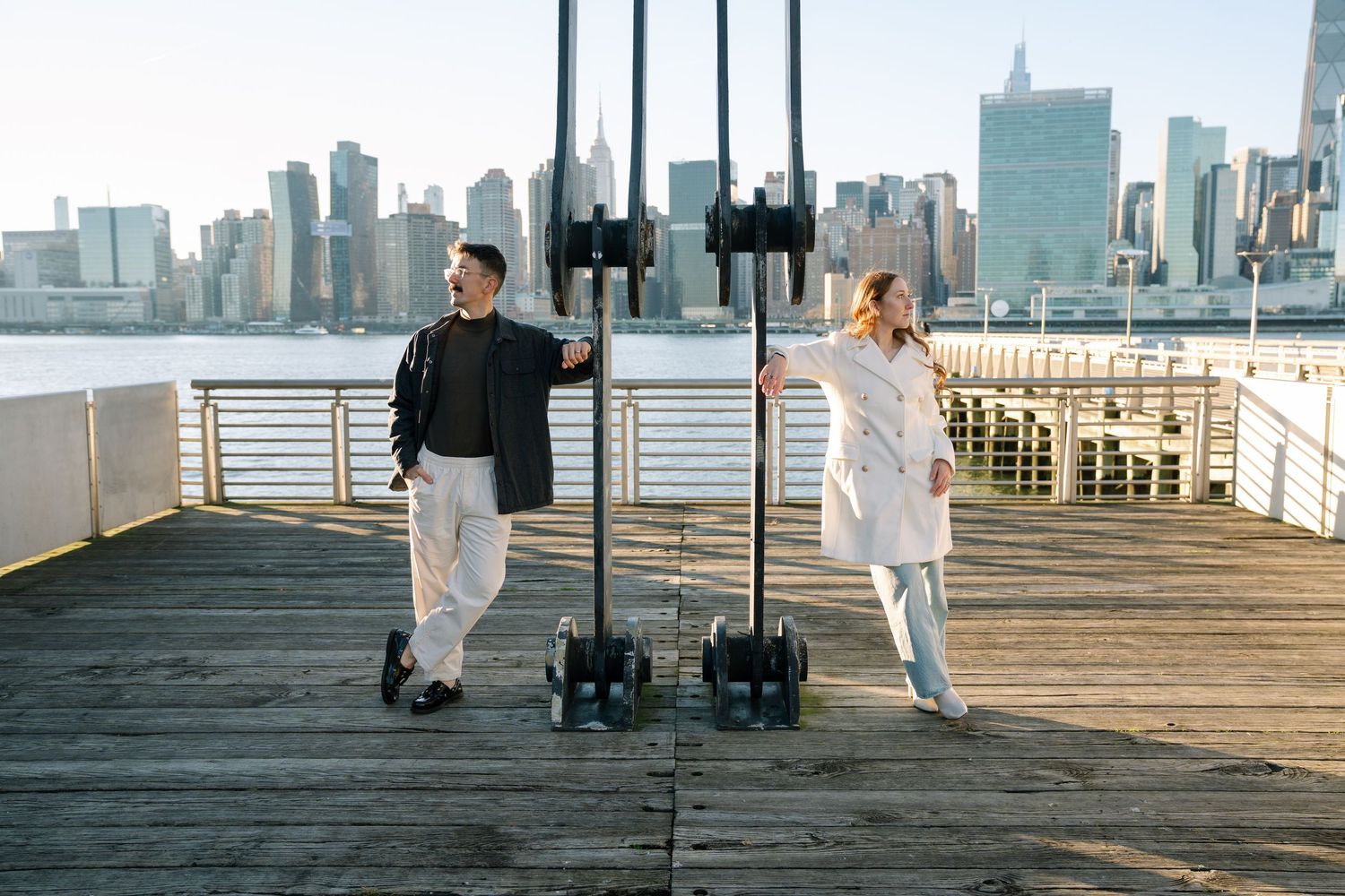 Couple walking together on a wooden pier with New York City skyline in the background during sunset.