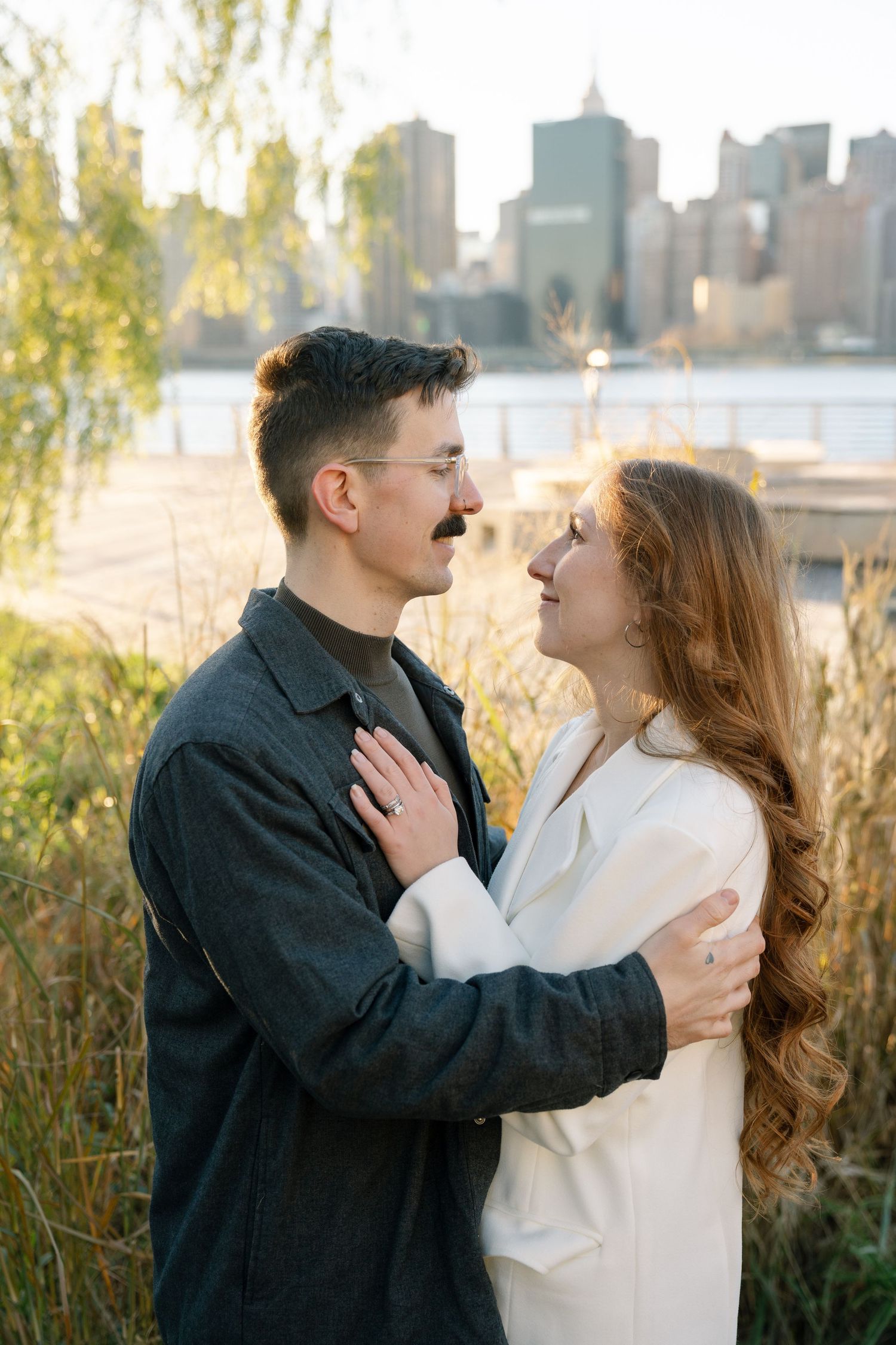 A romantic couple embraces by the waterfront with the Manhattan skyline visible in the golden hour light.
