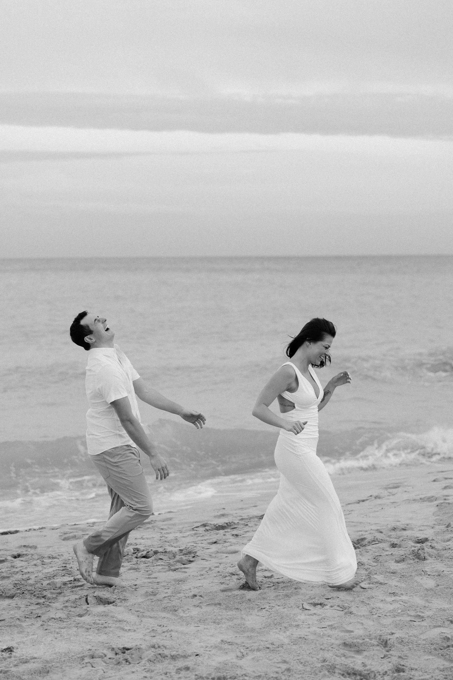 A playful couple falls dramatically on the beach in black and white photography.
