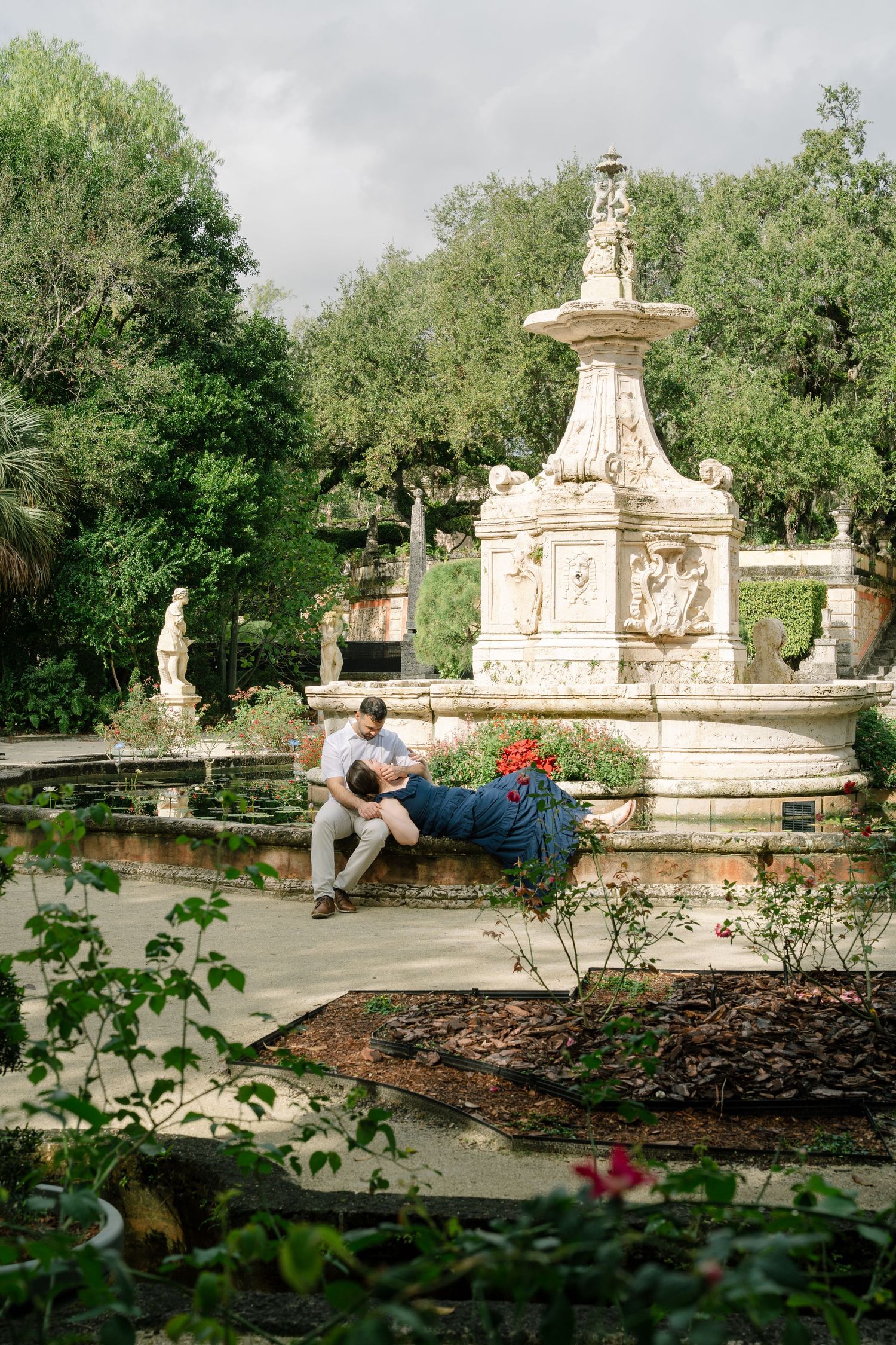 Ornate stone fountain and garden architecture in a classical style surrounded by lush greenery on a cloudy day.