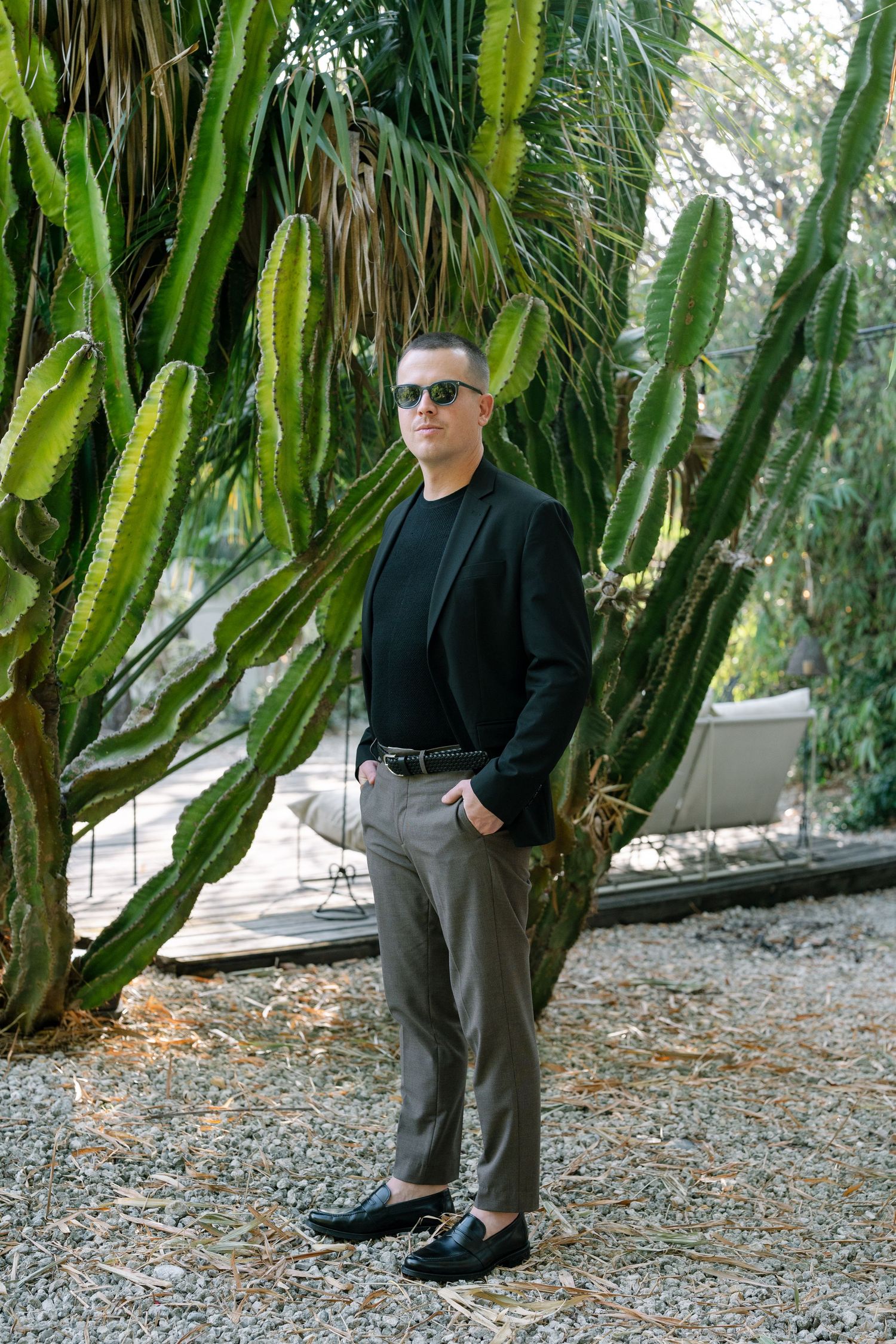 Person in black jacket and gray pants stands among tall cactus plants in garden setting.