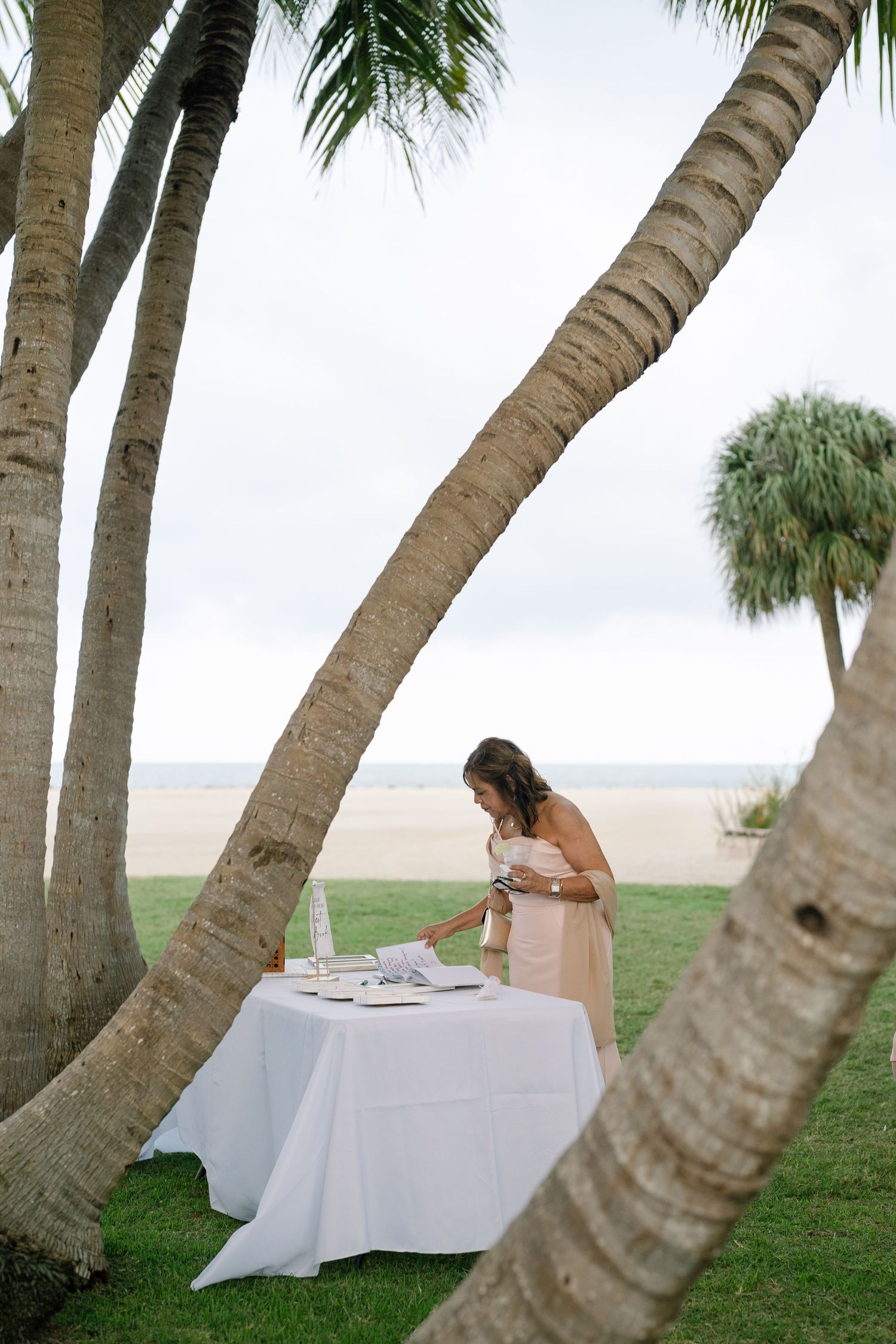 Bridesmaid in blush gown at guest book table framed by leaning palm trees on beach.