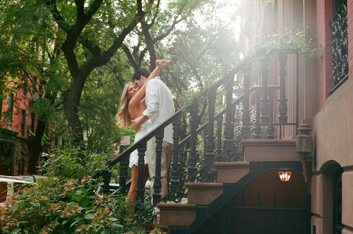 A romantic couple shares a kiss on a sunlit brownstone stoop surrounded by lush green trees in New York City.
