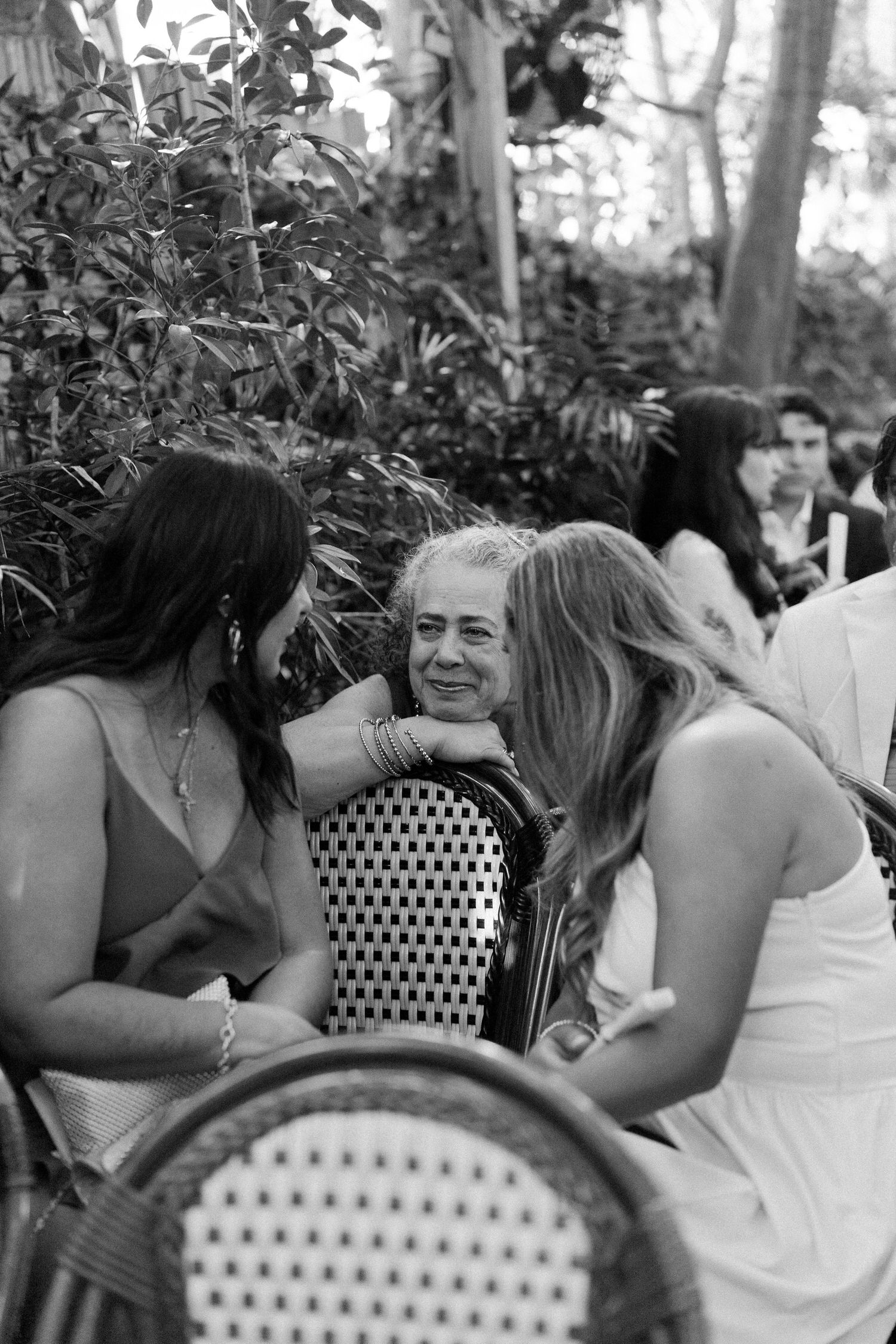 Black and white photo of three women chatting at an outdoor wedding reception surrounded by lush greenery.