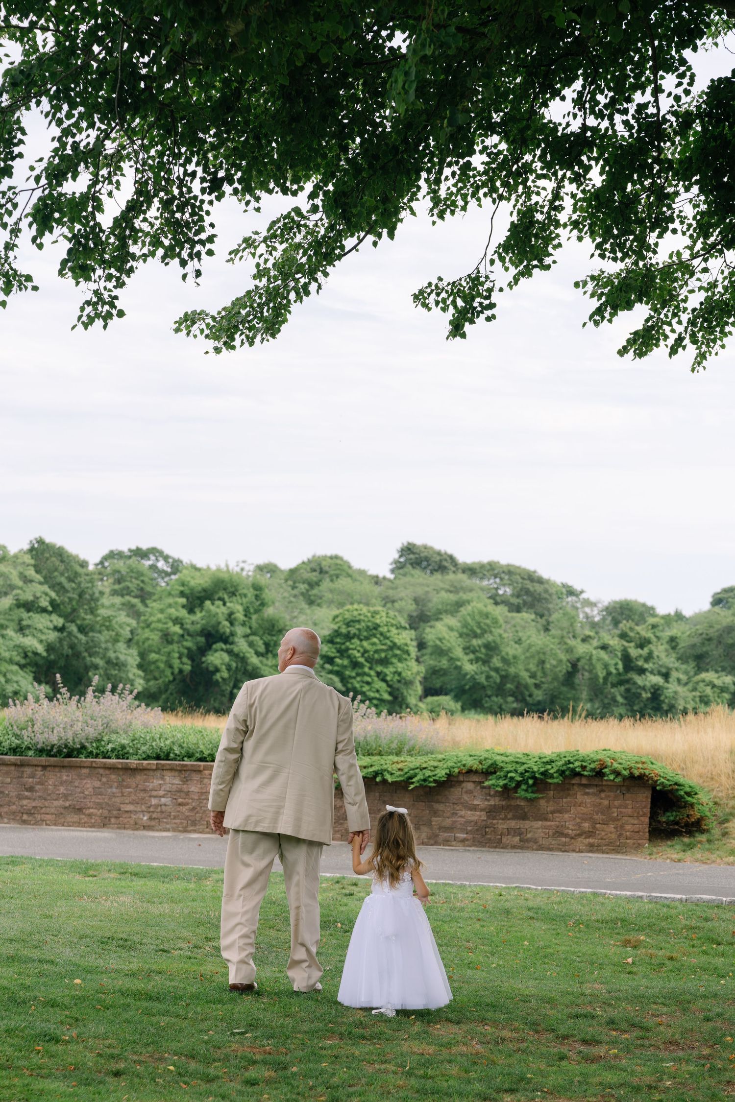 An elderly man holds hands with a young flower girl in a white dress, walking through a lush green park.