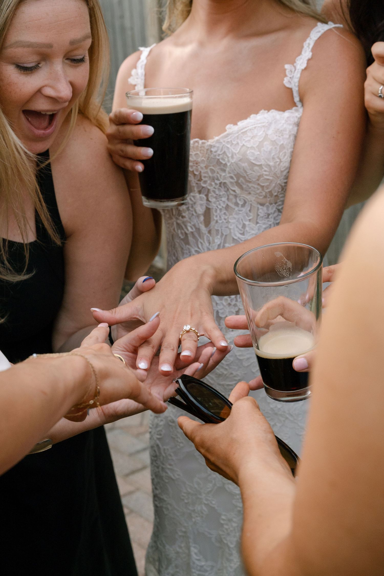 Wedding guests admire the brides ring while holding dark beer glasses at an outdoor wedding celebration.