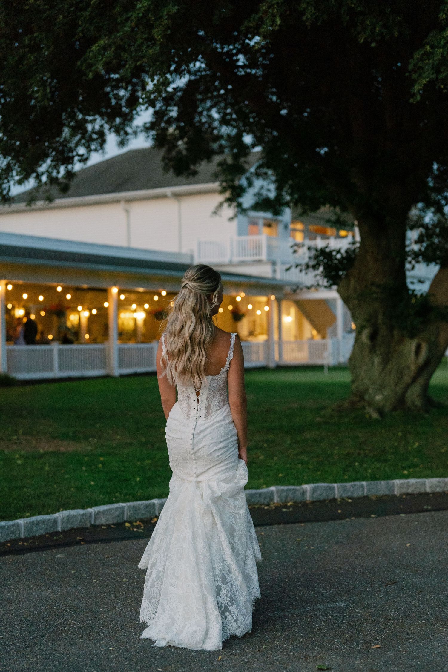 A bride in a fitted lace wedding gown stands outside a warmly lit venue at dusk.