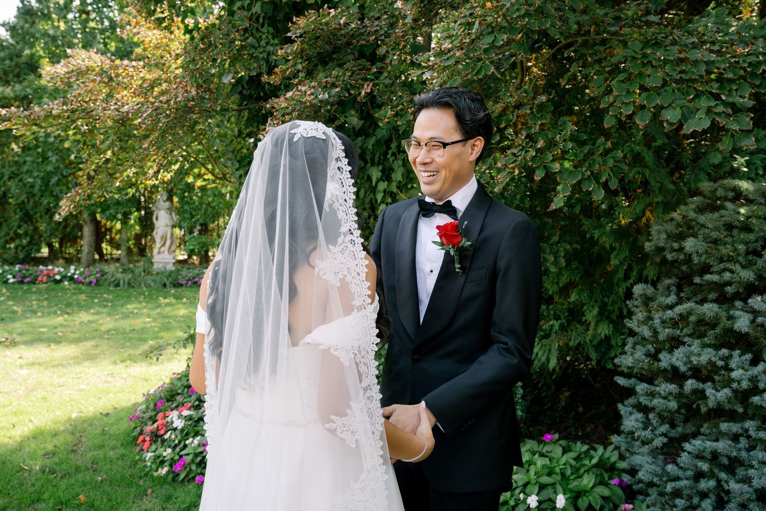A couple in wedding attire poses outdoors in a lush garden setting with green foliage in the background.
