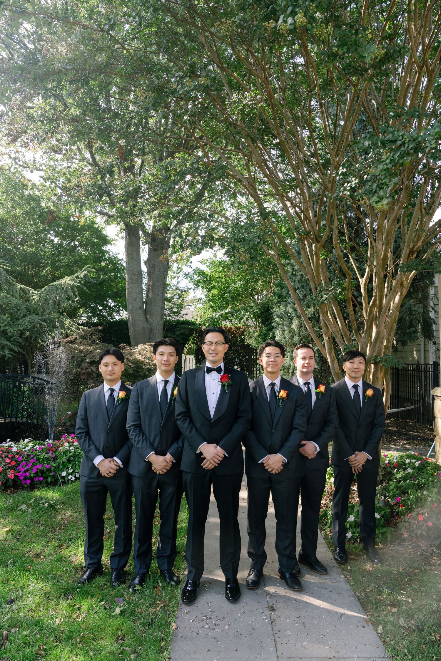 Wedding party in formal black suits posing together on a garden path with trees and flowers in the background.