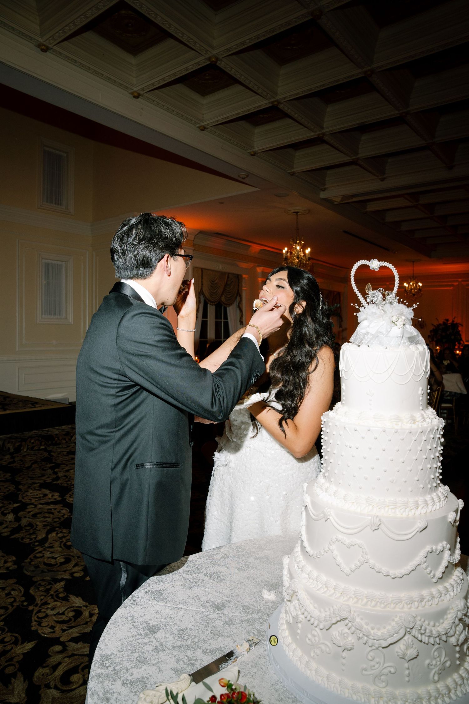 A couple shares cake during their wedding reception beside an elegant white tiered cake.