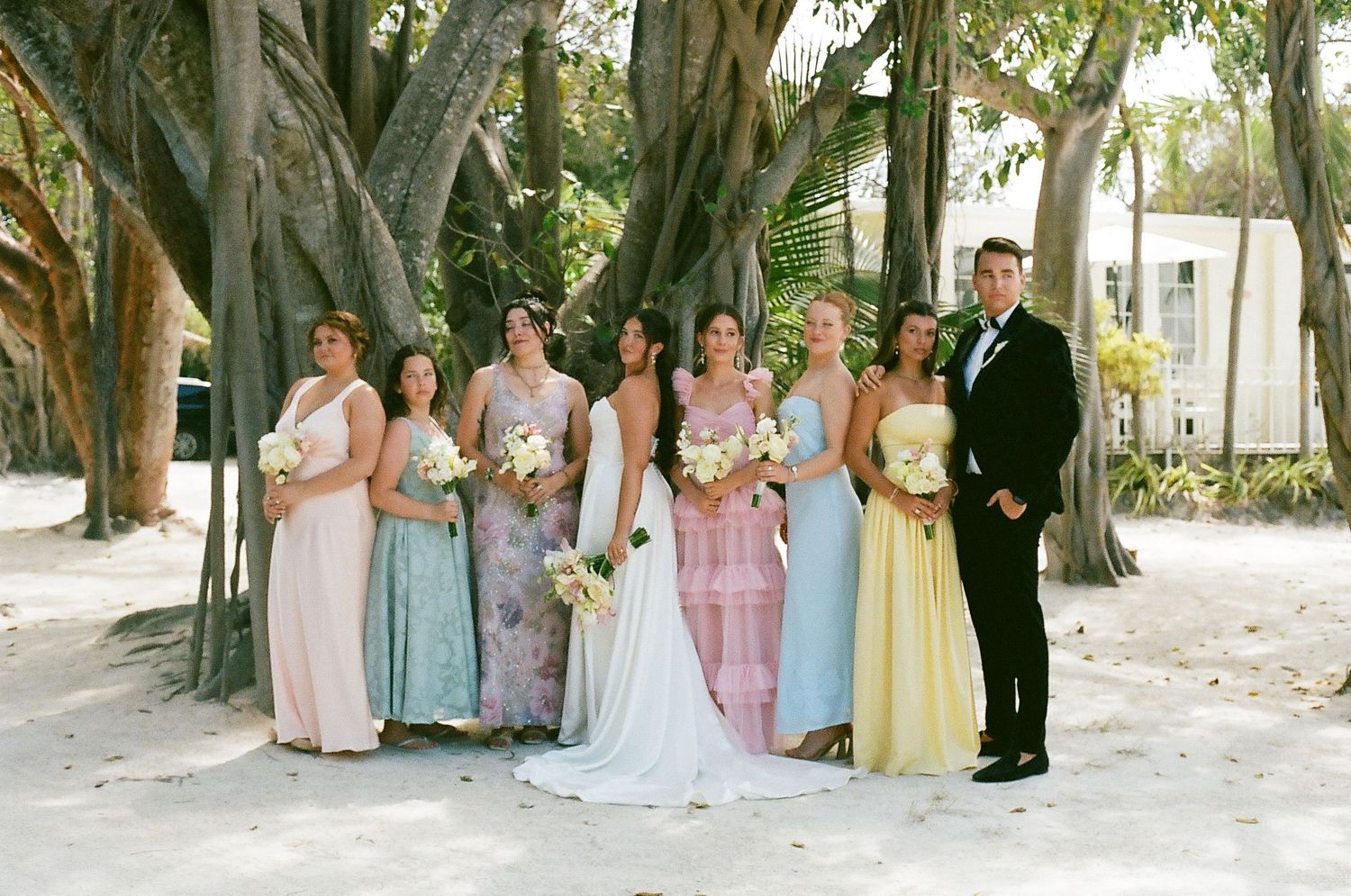 Bride and bridesmaids in pastel dresses pose with groom under banyan trees on sandy beach at tropical wedding.