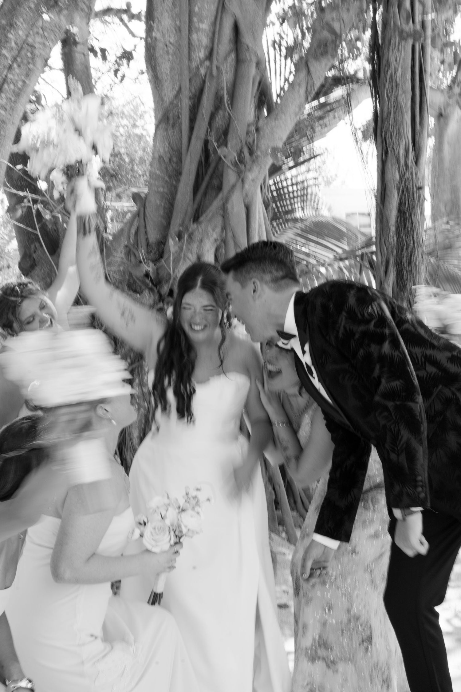 Black and white wedding photo of smiling bride in white gown with groom in suit celebrating outdoors under trees.