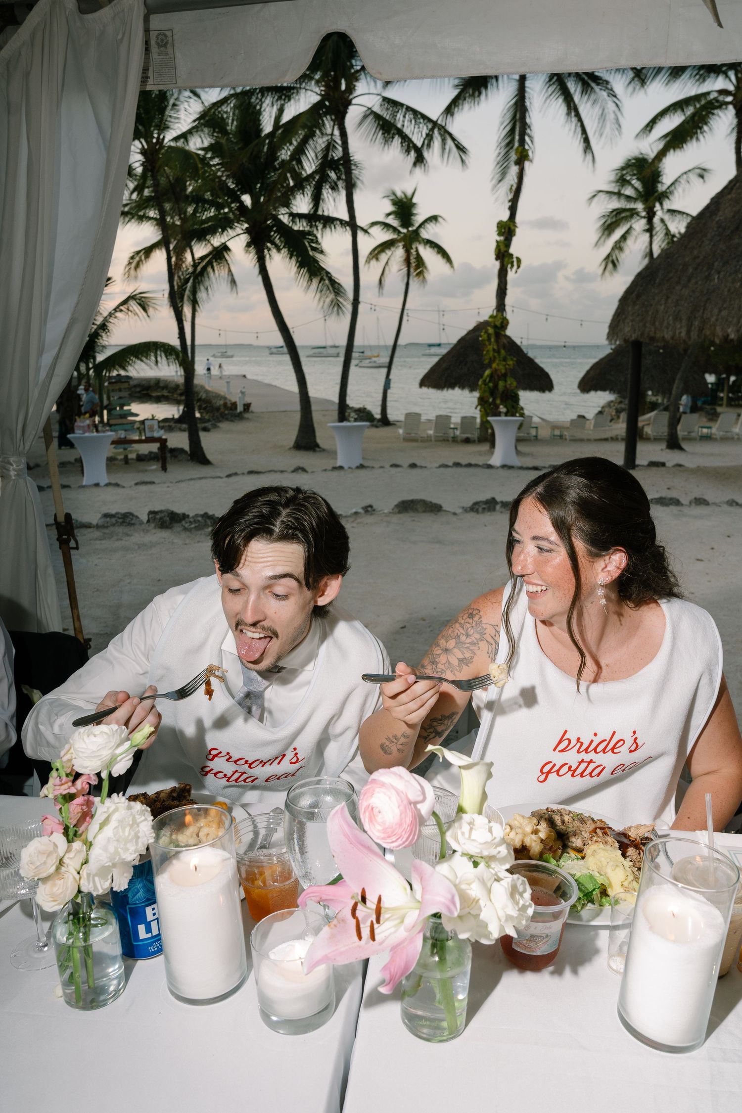 A couple in wedding attire sit at a table with flowers and drinks on a beach.