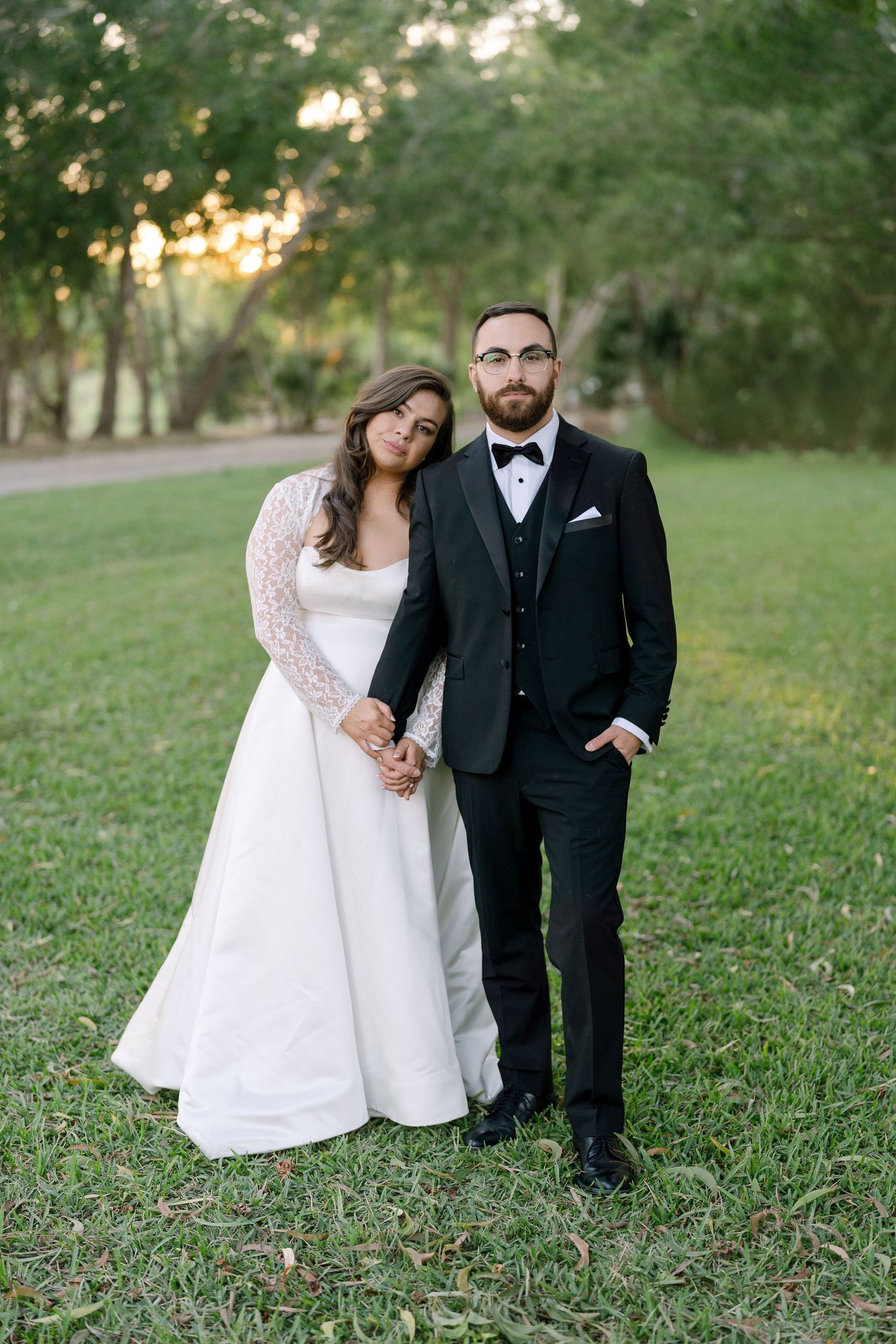A bride and groom stand together in a grassy field, with the bride wearing a white dress and the groom wearing a black suit.