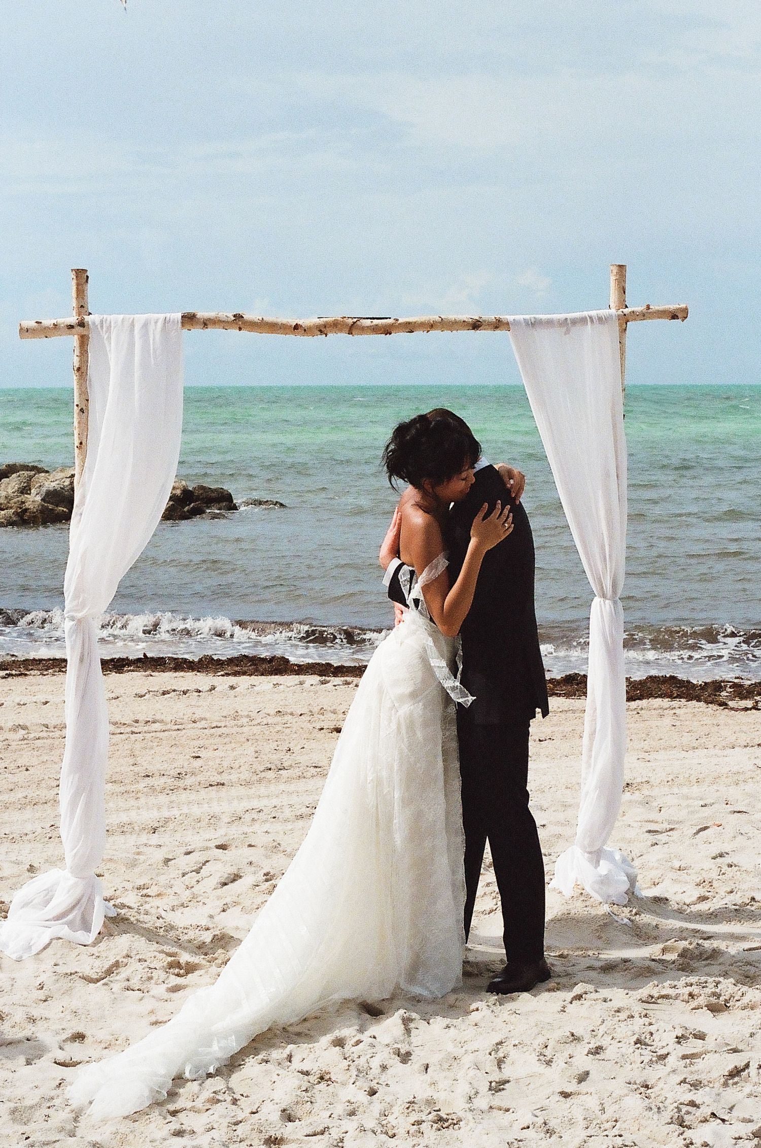 Couple sharing first kiss under white draped arch during romantic beach wedding ceremony with turquoise ocean backdrop.
