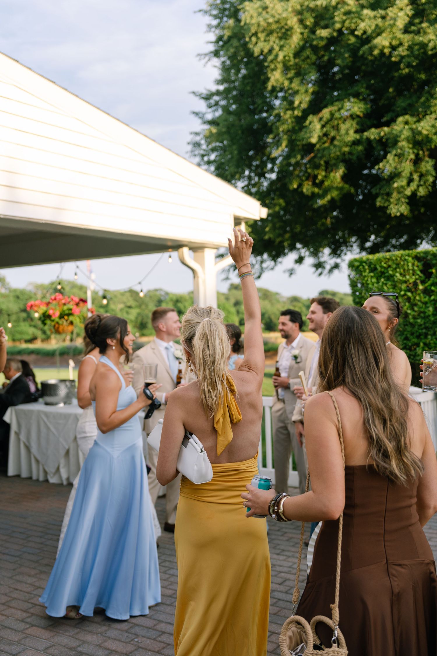 Guests in elegant dresses mingle outdoors at a summer wedding reception under string lights.