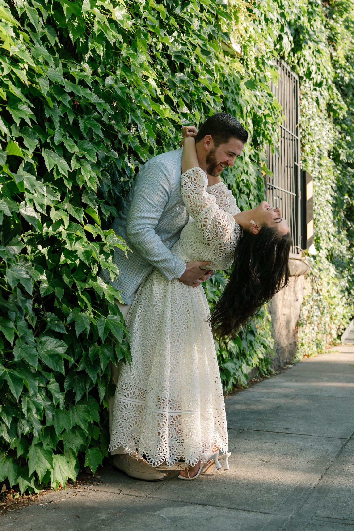 A couple dancing and laughing in front of a lush ivy-covered wall, woman in white lace dress.