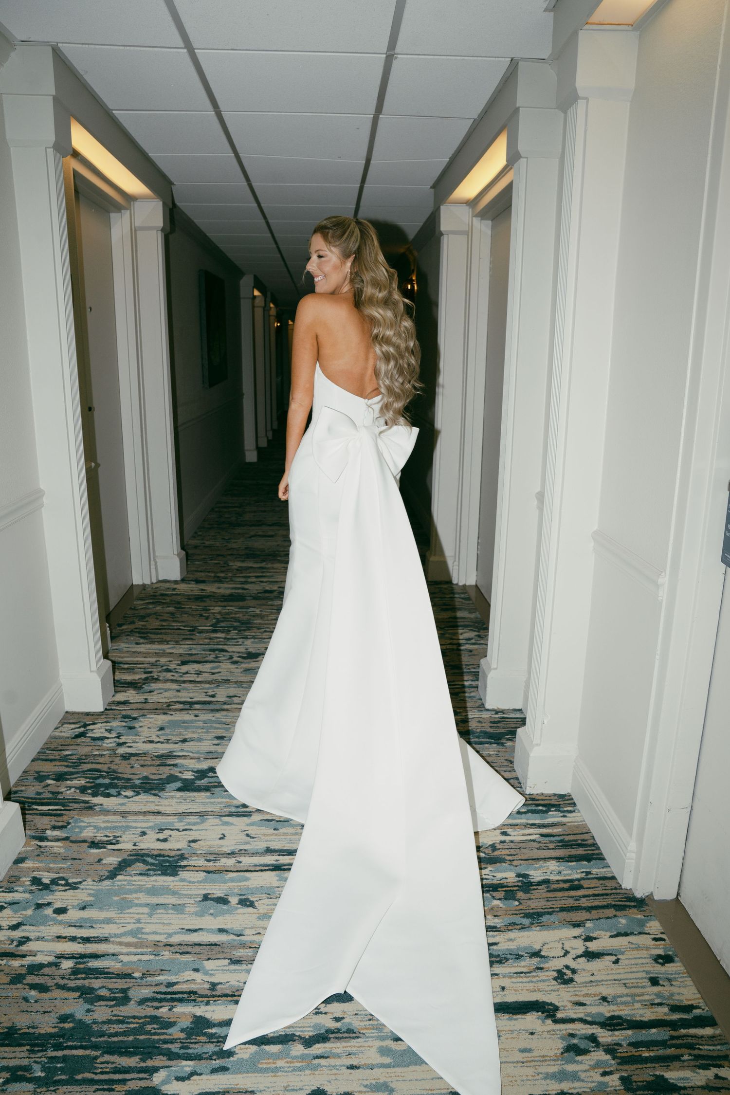 Back view of a bride in a flowing white wedding gown with dramatic train standing in a hotel hallway.
