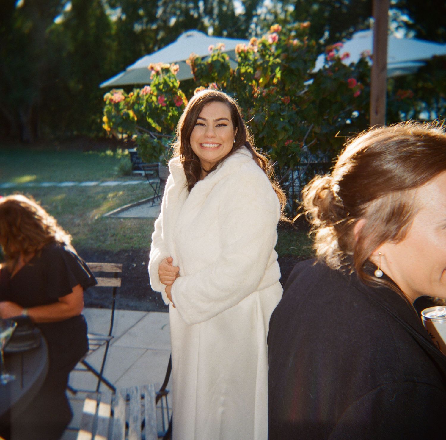 Someone in a cozy white robe smiles at an outdoor gathering with string lights and people in the background.