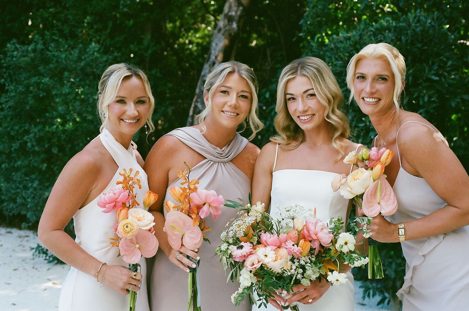 Row of bridesmaids in matching white dresses posing outdoors with peach and pink bouquets.