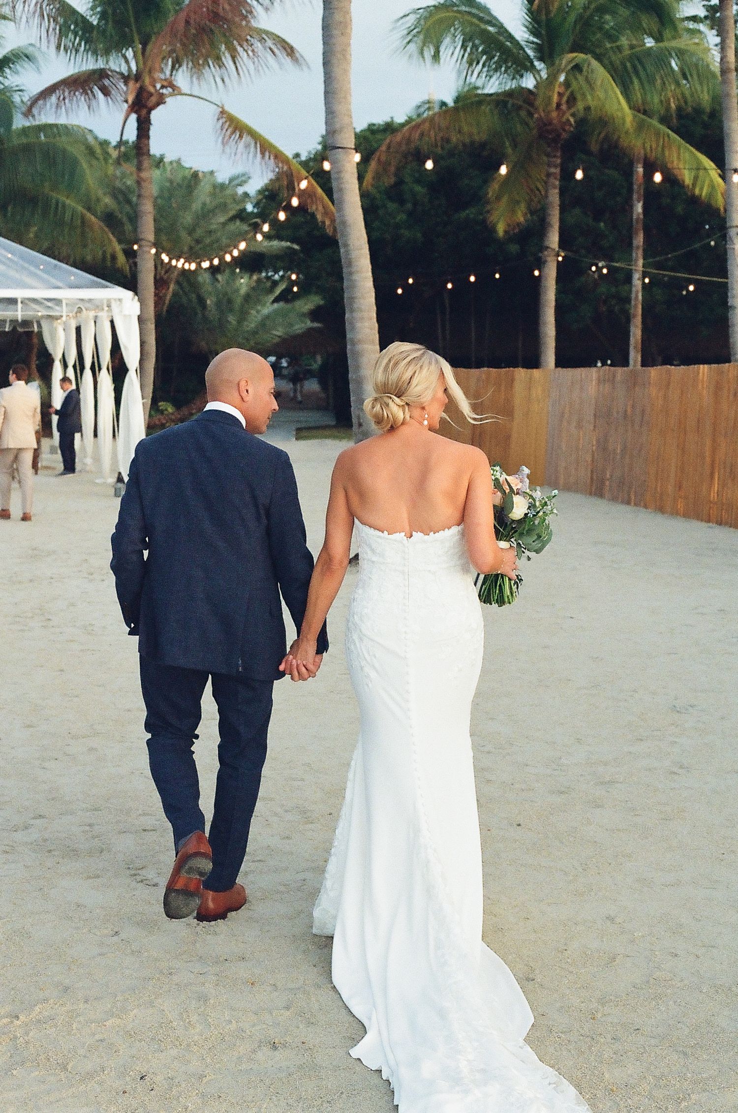 Wedding party walking along a sandy beach path with palm trees and string lights.