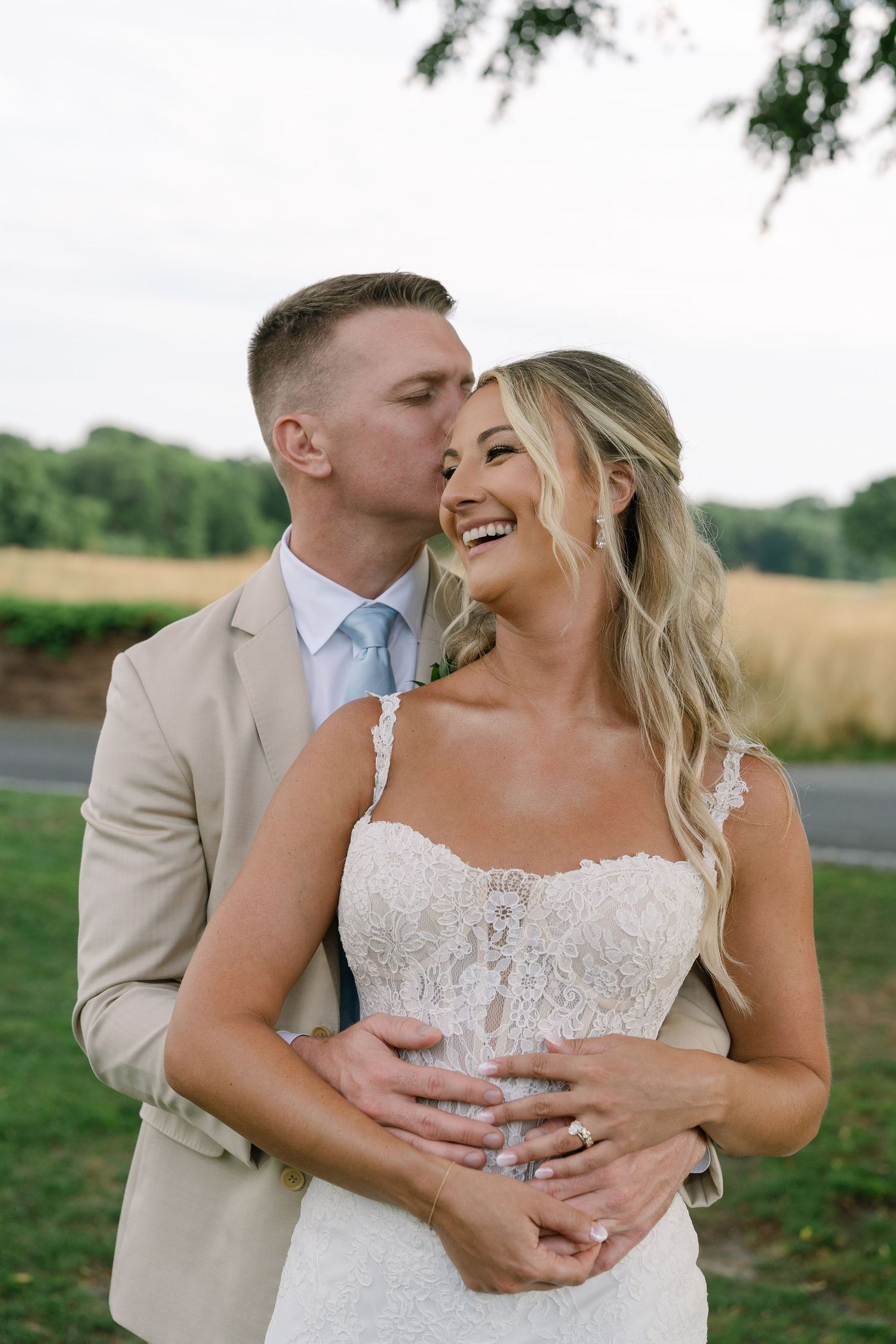 Bride in lace gown laughs joyfully as groom in tan suit kisses her cheek outdoors at golden hour.