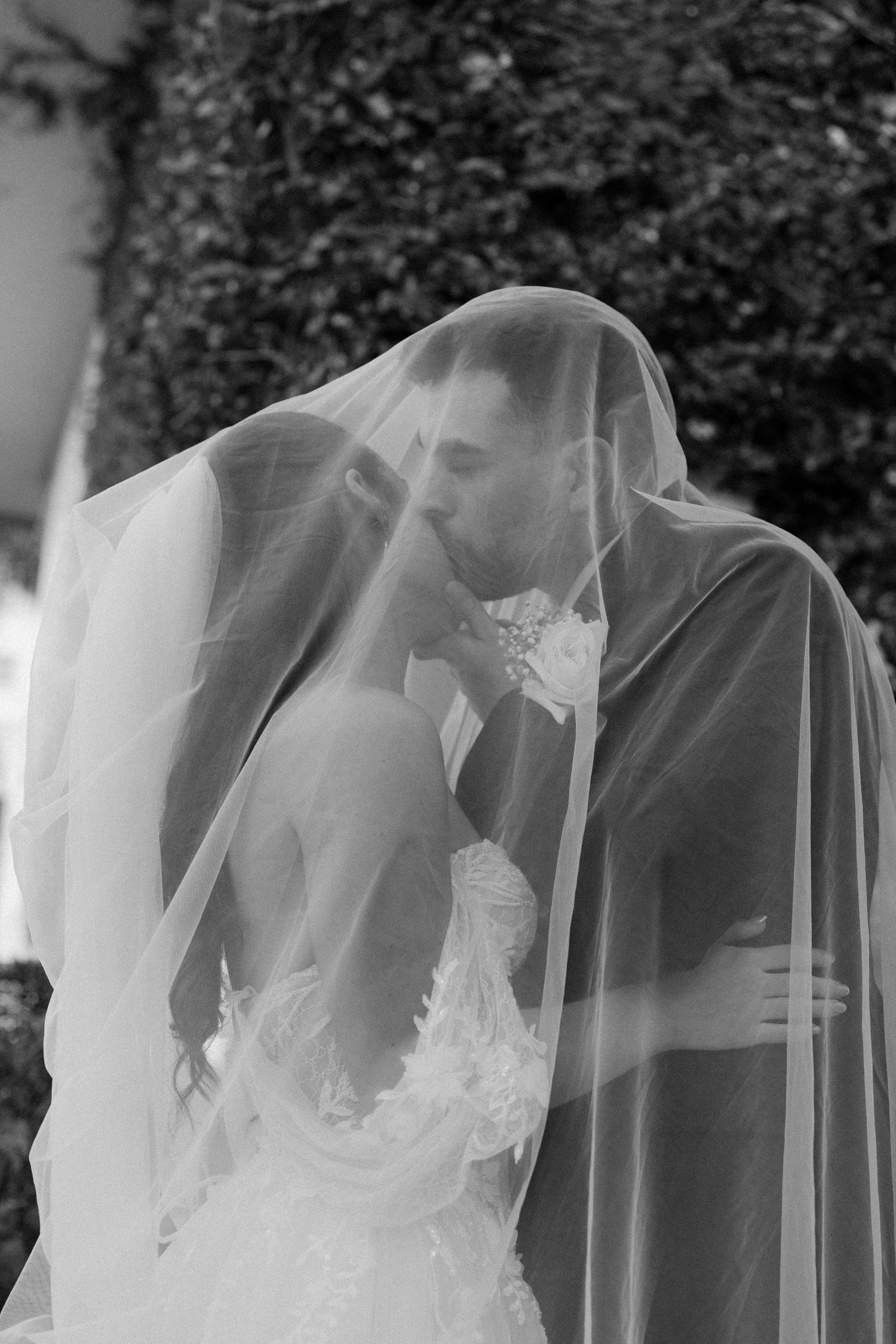 Bride and groom share a romantic kiss under a flowing white veil in a black and white wedding portrait.