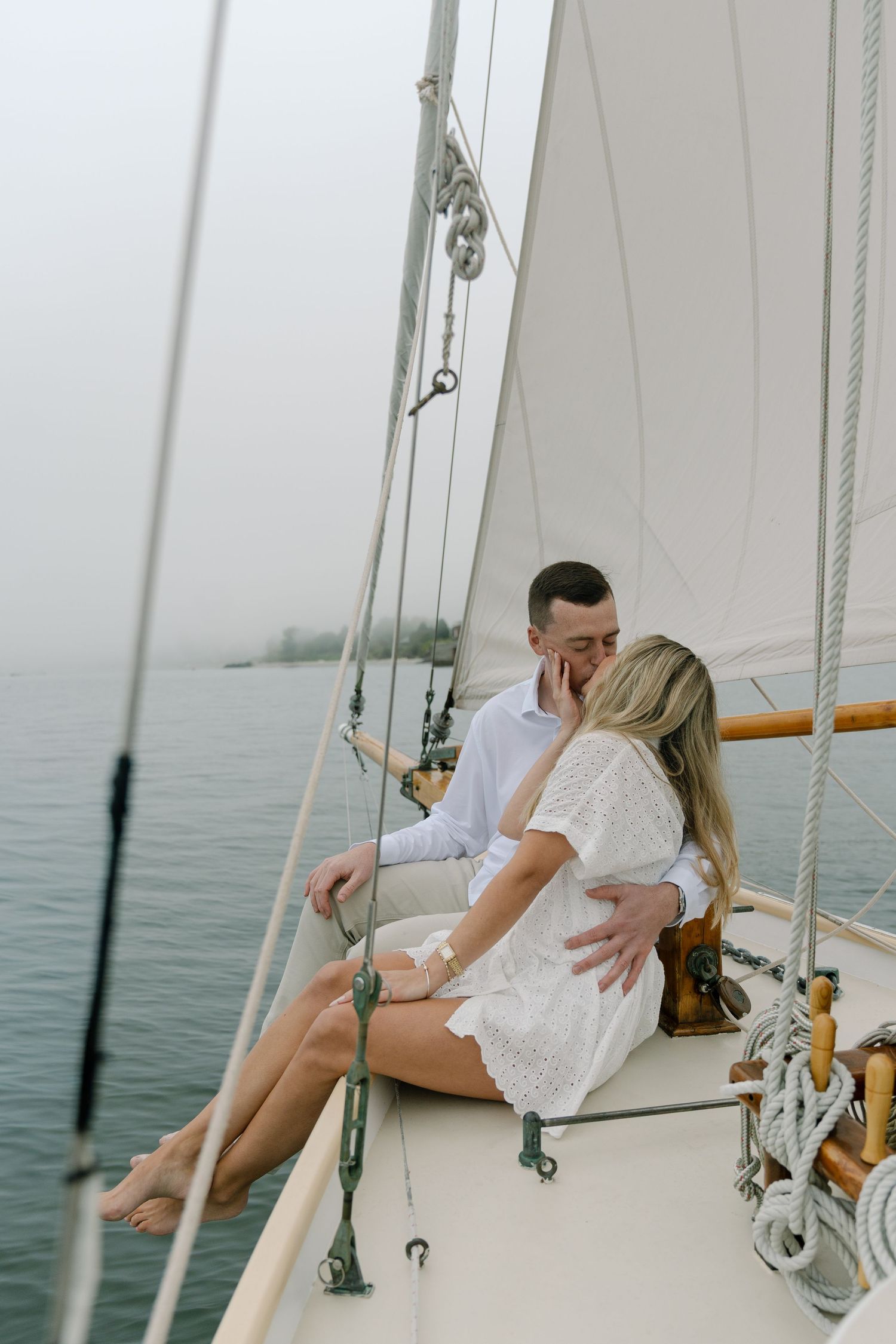 Couple sharing a romantic kiss on a sailboat deck, dressed in white, surrounded by misty water and white sails.