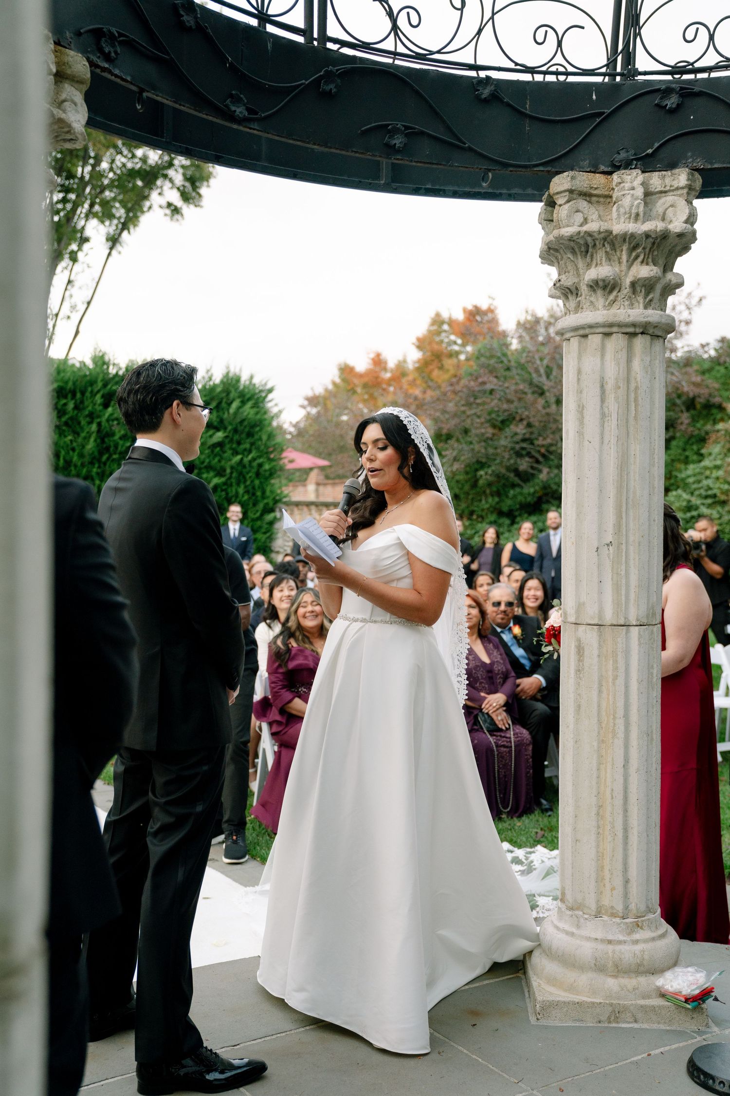 Wedding party gathered under an ornate gazebo with classical columns during an outdoor ceremony.