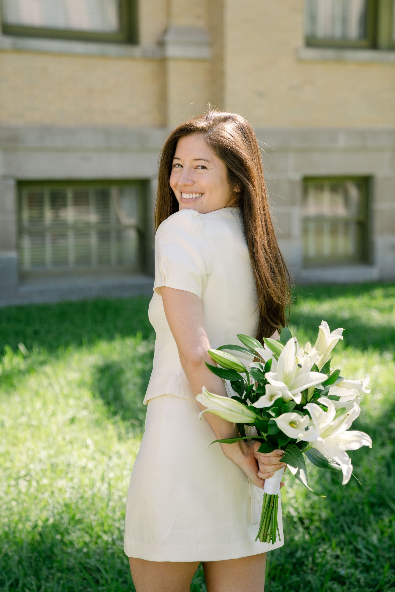 A person in a white dress holds a bouquet of elegant white lilies while standing on a sunny lawn.