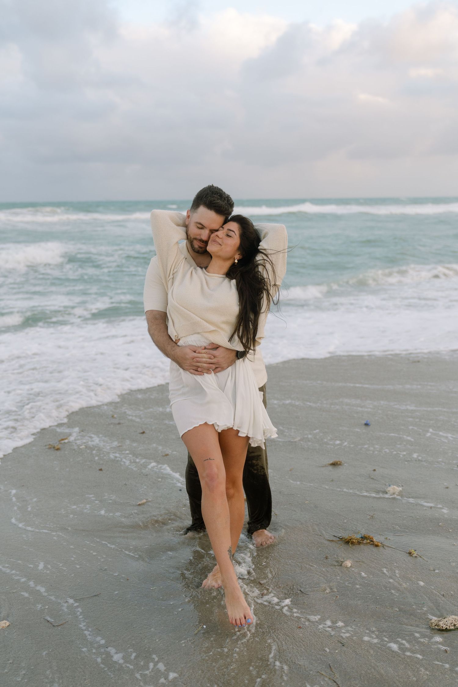 A romantic couple shares tender moments on a cloudy beach as waves crash behind them.