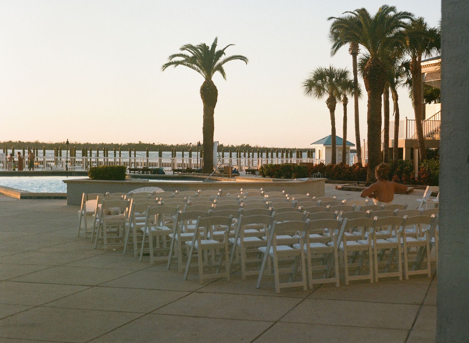 Empty white chairs arranged in rows on a beach with palm trees and a pier silhouetted against the sunset.