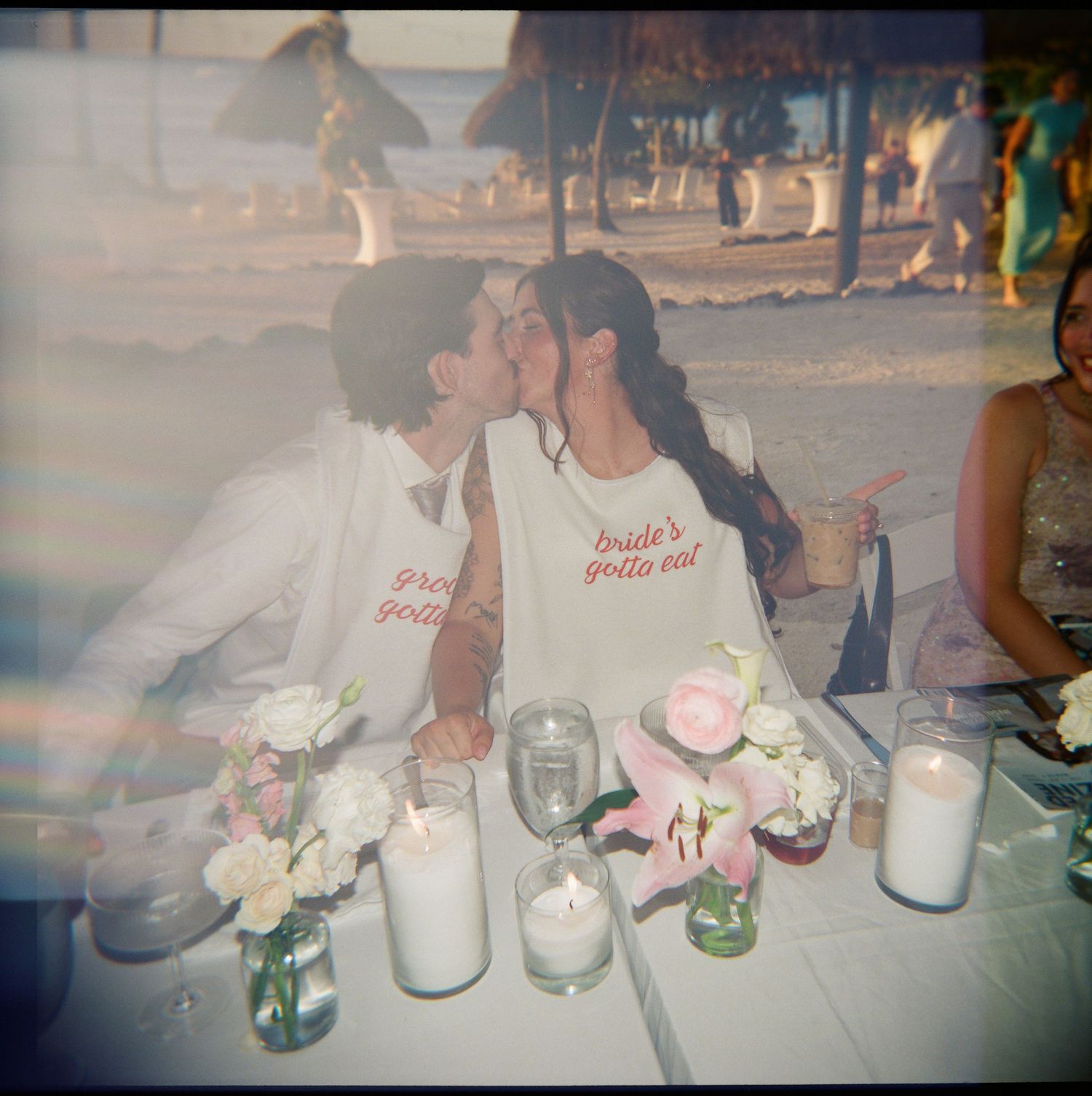 Newlyweds kissing at reception table wearing matching white aprons at beach wedding dinner.