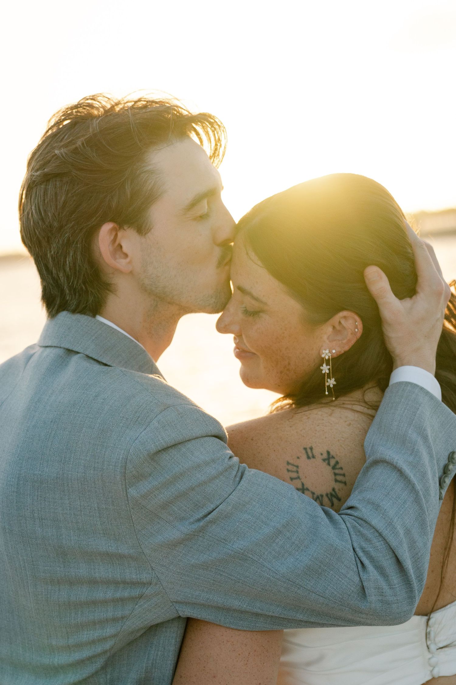 A bride and groom embracing and kissing in an outdoor setting with the sun shining on them.