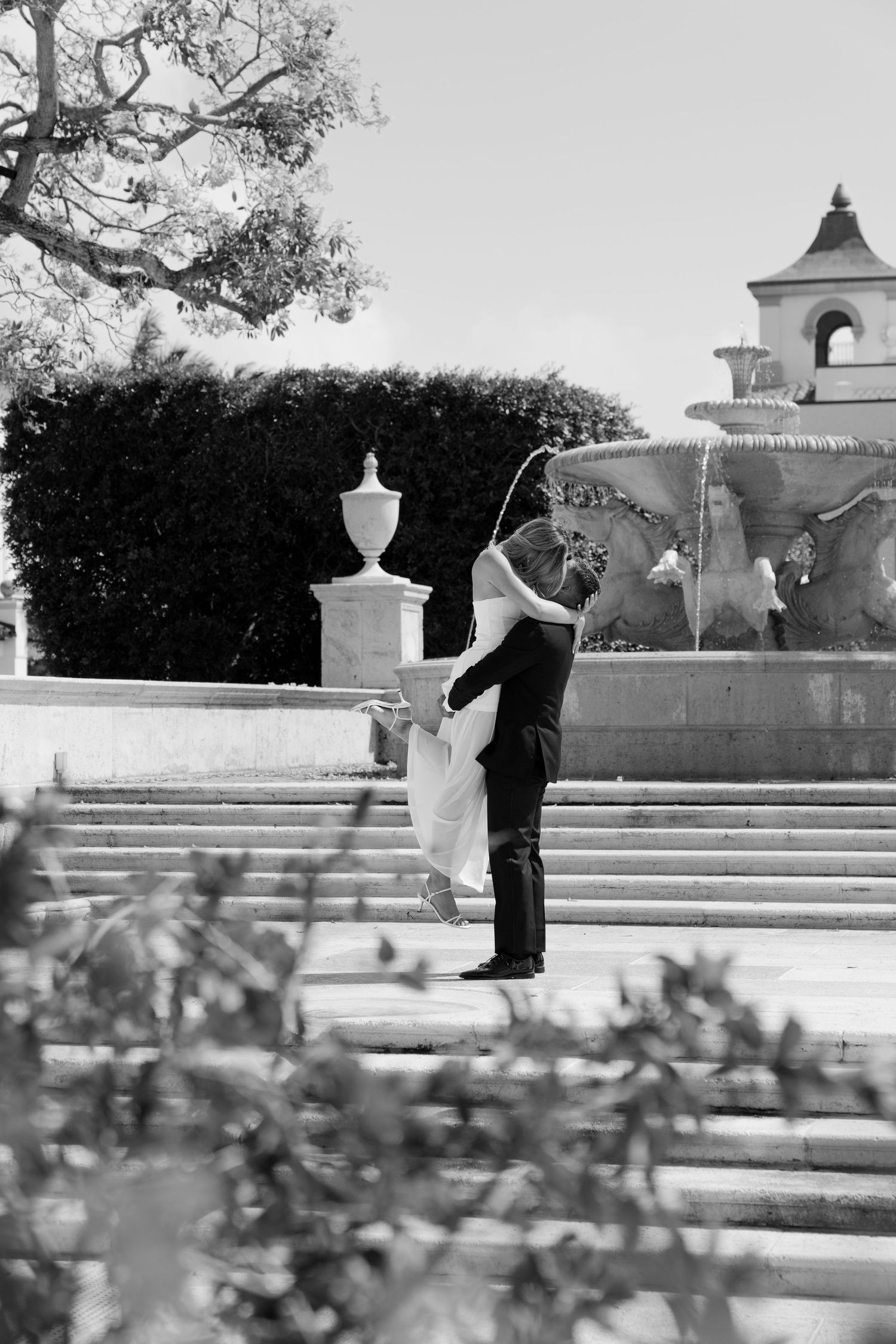 Black and white photo of a couple embracing on steps near a fountain at an elegant outdoor wedding venue.