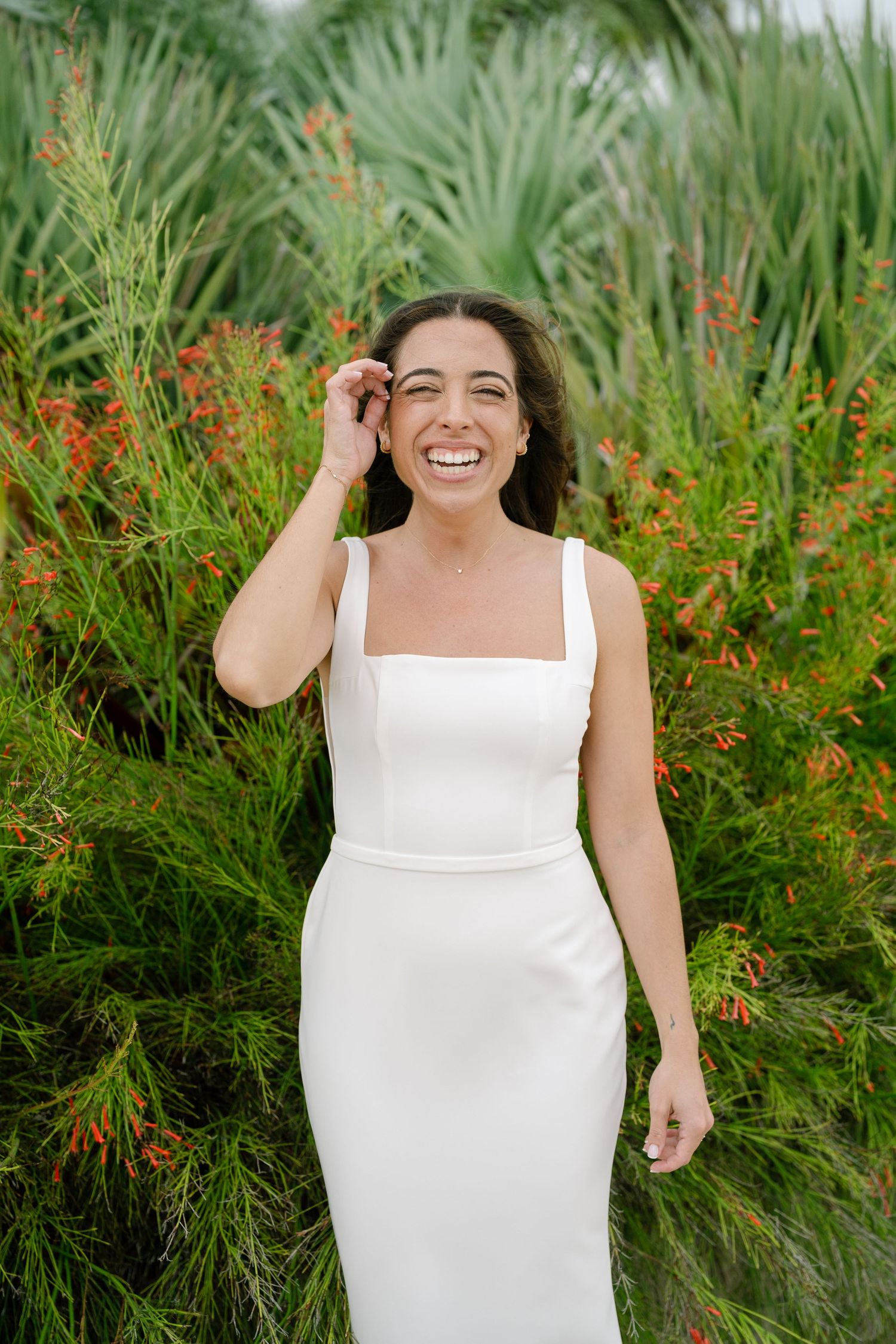 Smiling bride in white sleeveless gown standing among lush green tropical plants and red flowers.