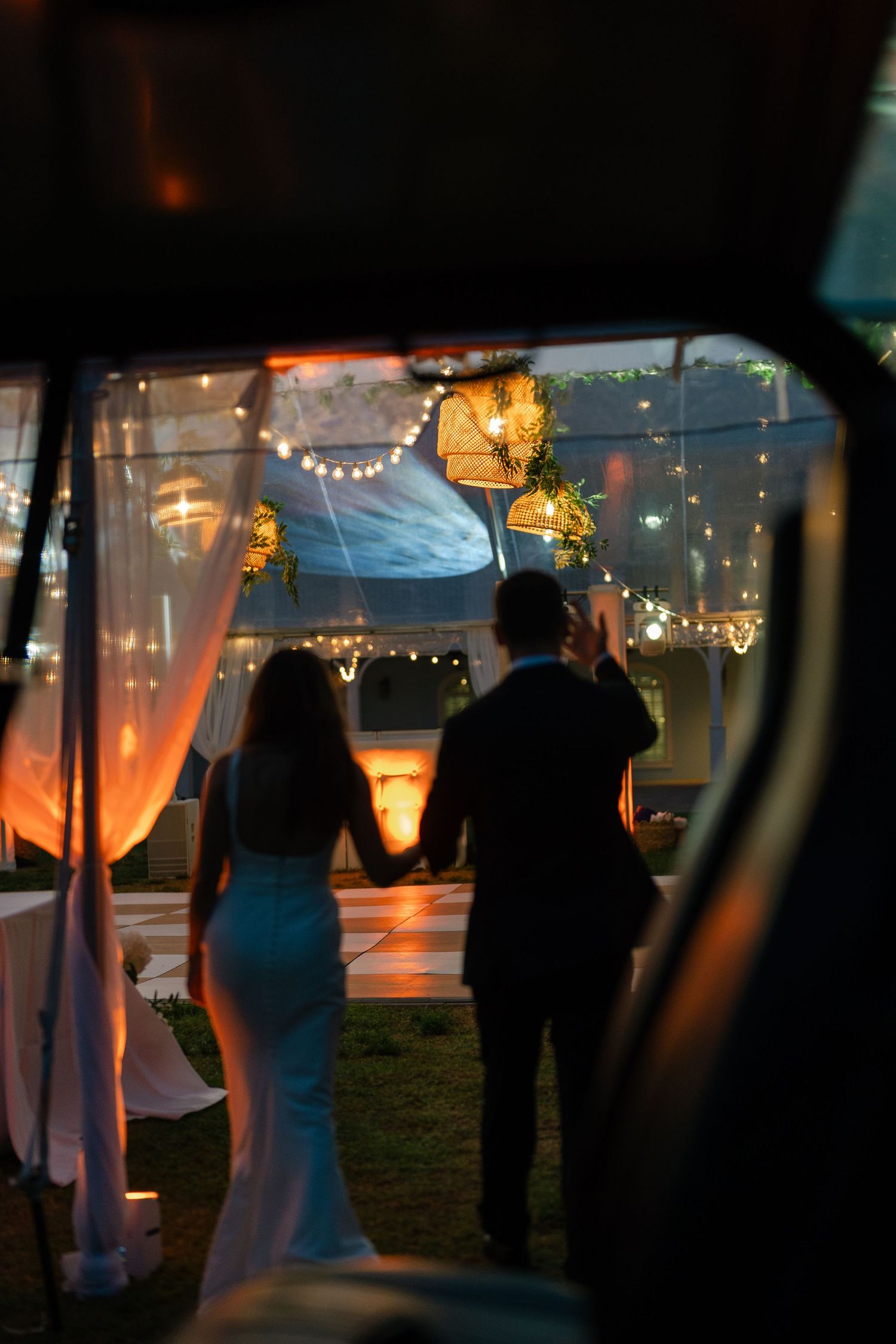 Silhouette of bride and groom entering lit wedding reception tent with string lights at night.